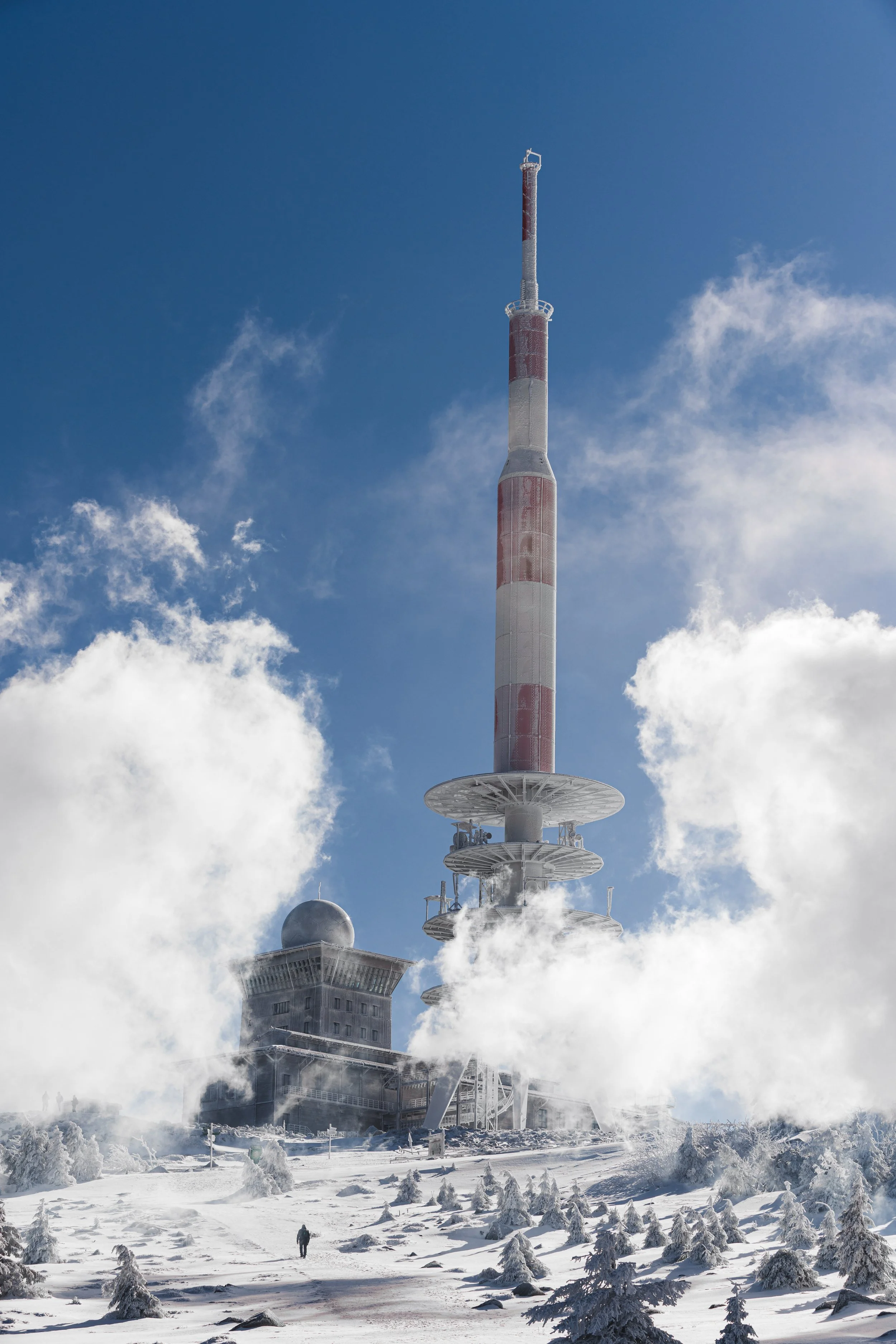 Ein hoher Fernsehturm auf einer verschneiten Berglandschaft mit vielen kleinen verschneiten Bäumen, Wolken und einer Person im Vordergrund.
