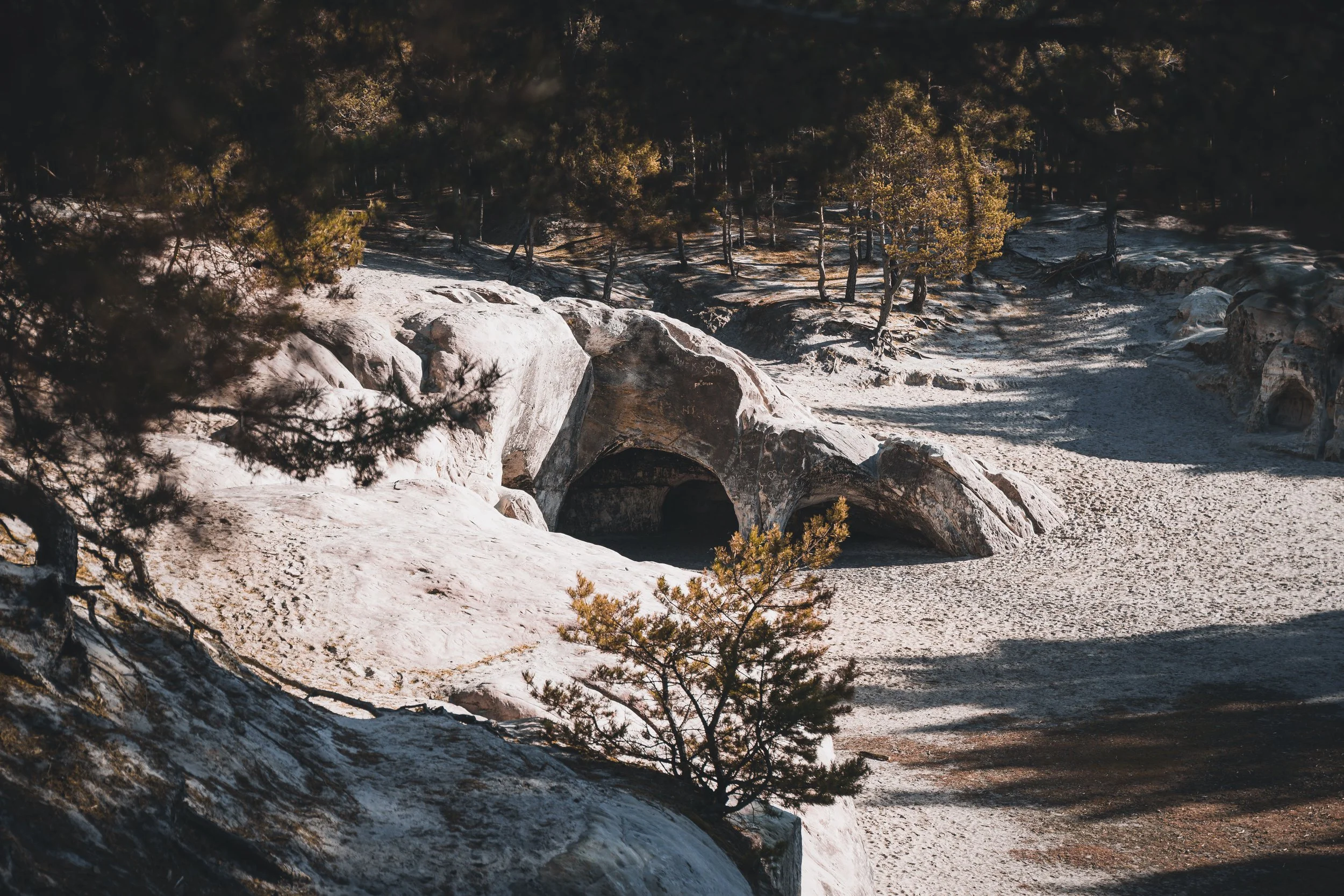 Felsformationen in einer trockenen, bewaldeten Landschaft mit Bäumen und Schatten, bei Sonnenlicht.