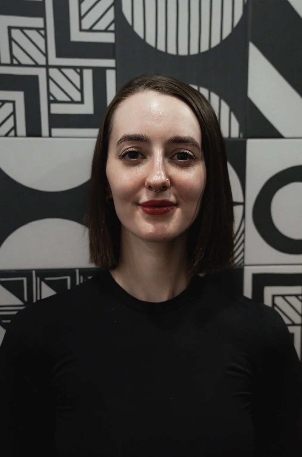 Person with long brown hair and a septum piercing, smiling in front of a white brick wall.