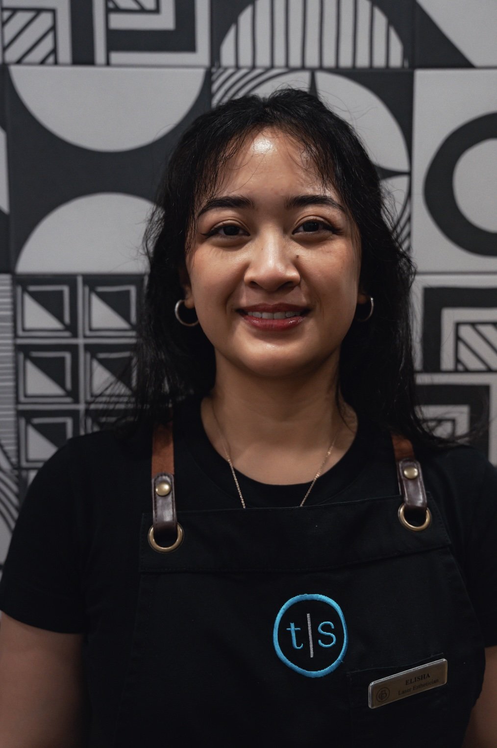 Person smiling in front of a white brick wall, wearing a black turtleneck and a necklace.