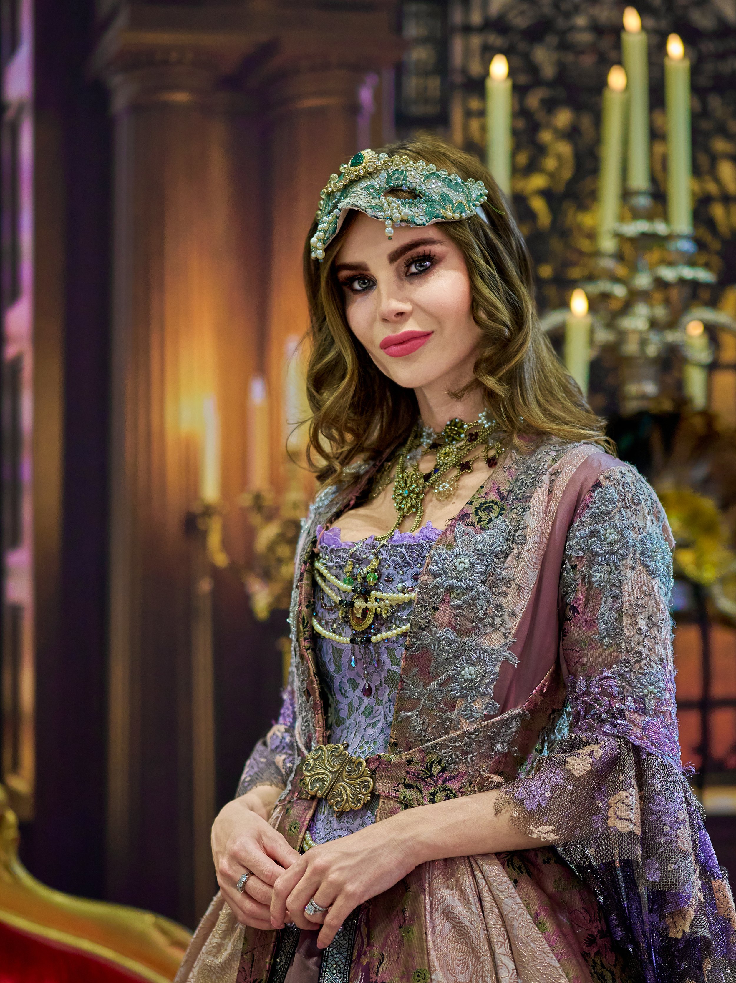 Woman in elaborate historical costume with beaded headdress and ornate necklace, standing in an opulent room with candles in the background.