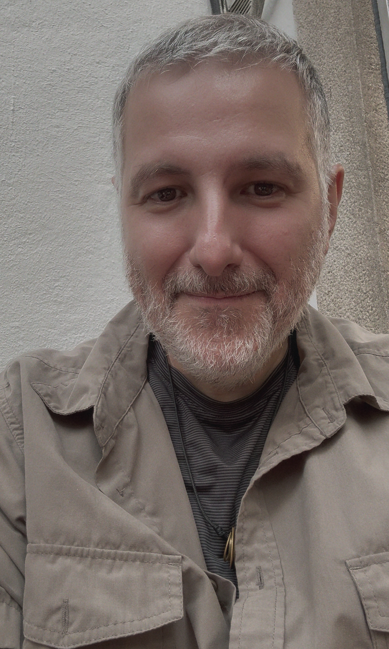 A smiling man with gray hair and beard taking a selfie outdoors against a beige wall.