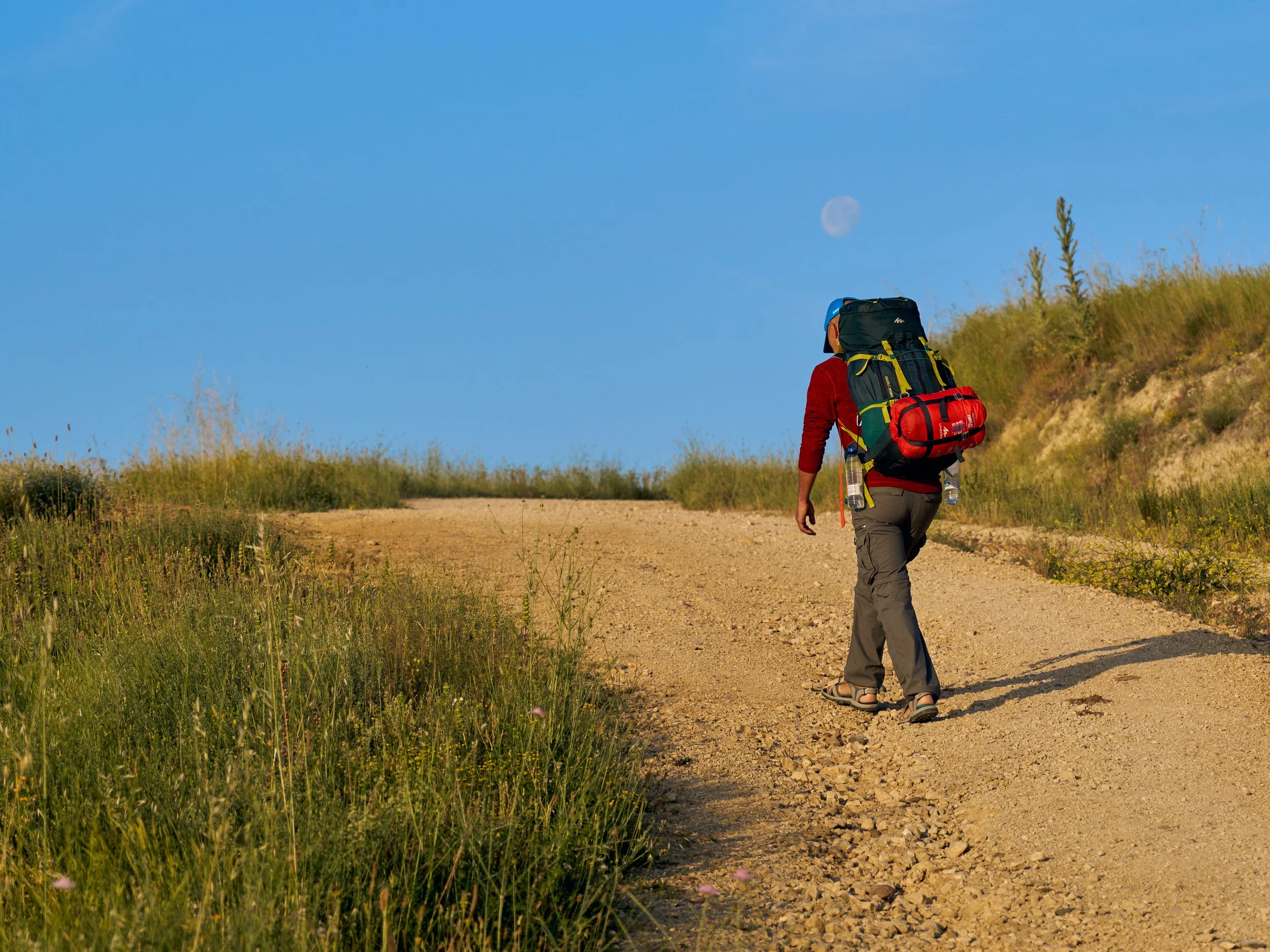 A person hiking on a dirt trail with a large backpack during daytime, with the moon visible in a clear blue sky and grassy hills on either side of the trail.
