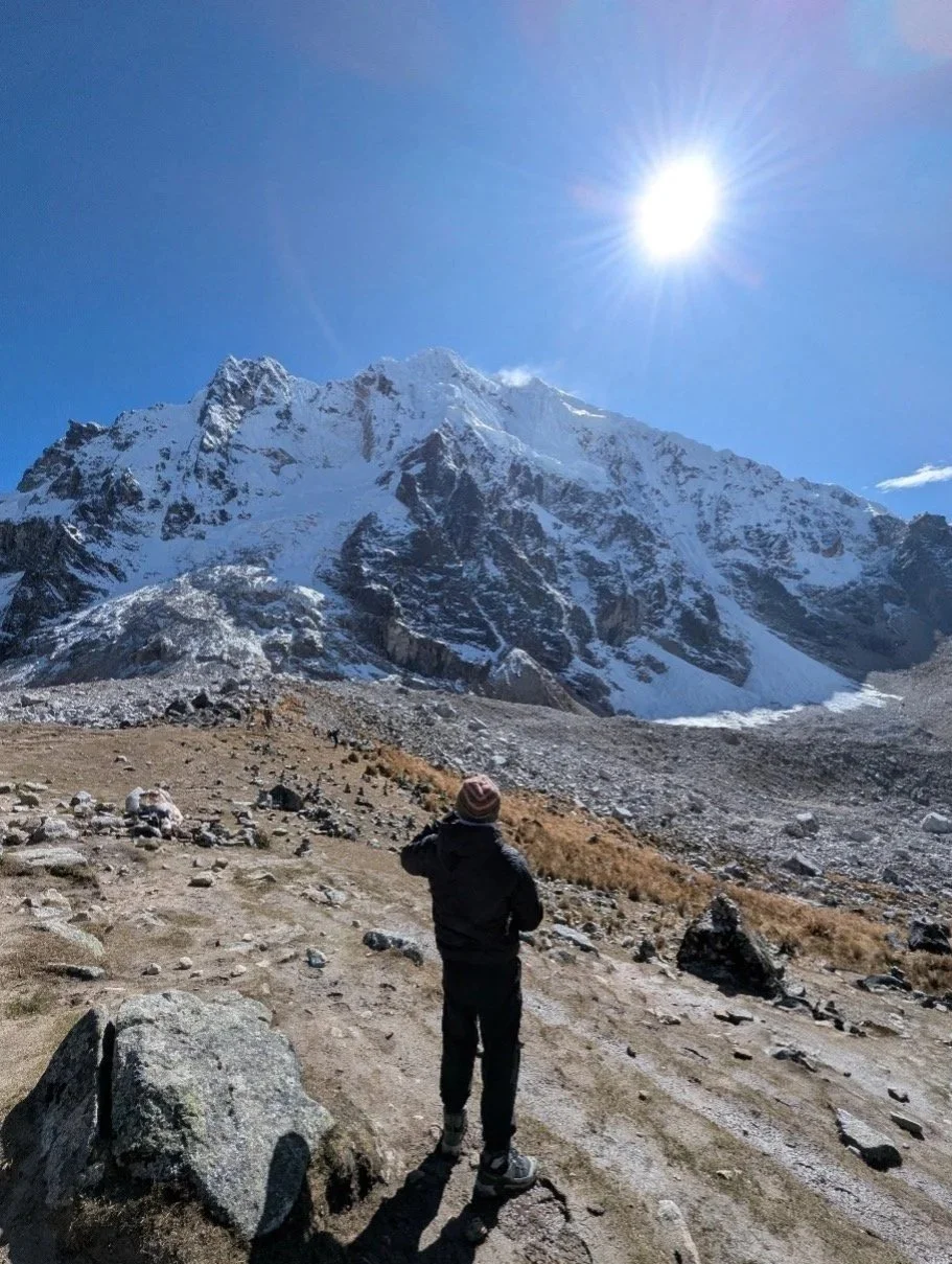 A person in dark clothing and a striped hat standing on a rocky, dirt trail in a mountainous landscape with snow-covered peaks under a bright sun in a clear blue sky.