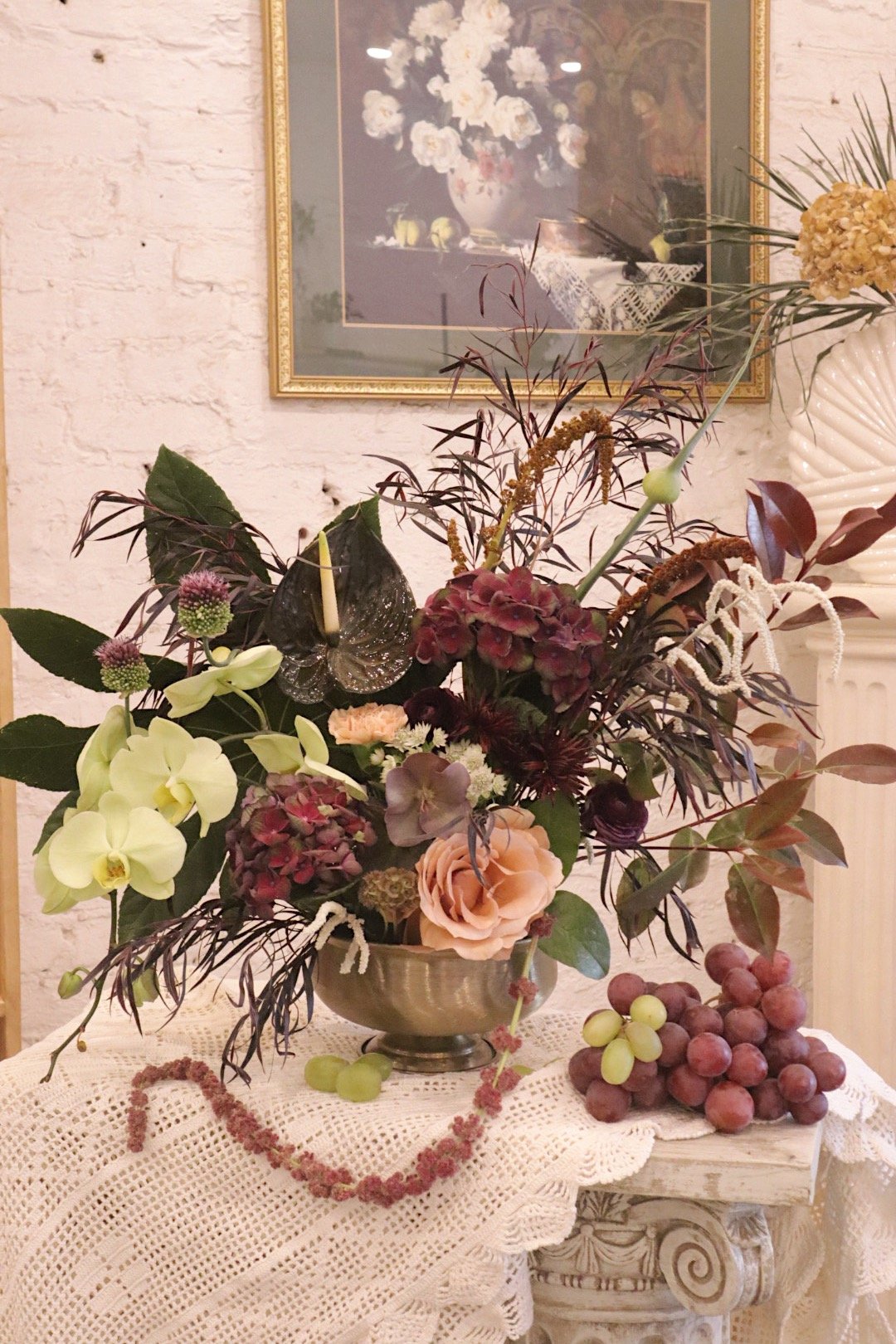 A floral arrangement with various flowers and greenery in a metallic bowl, set on a lace-covered table with grapes beside it. A framed painting of flowers hangs on a white brick wall in the background.