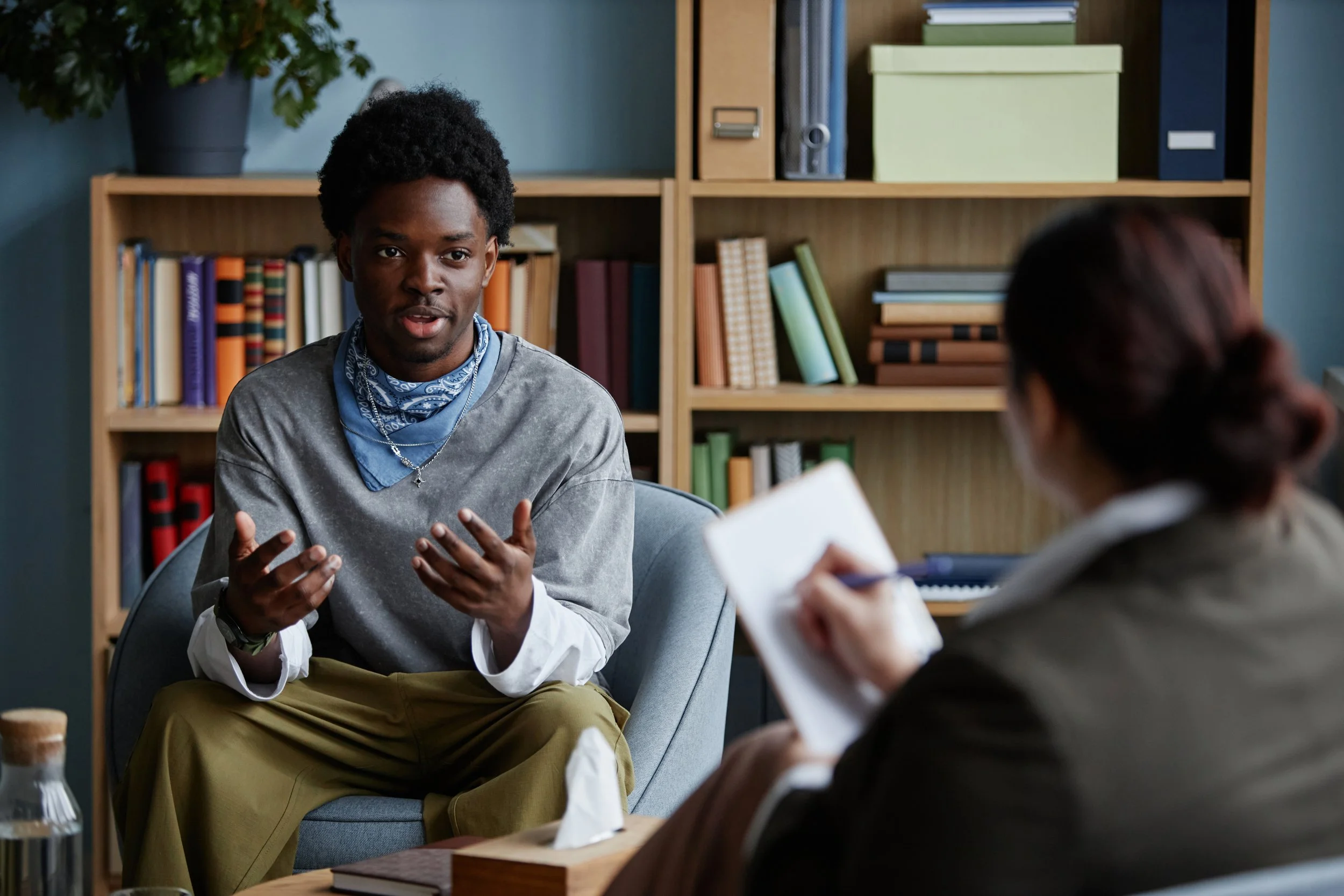 Black male sitting and talking with adult woman counselor during counseling session, gesturing with hands while woman taking notes, bookshelf with books in background