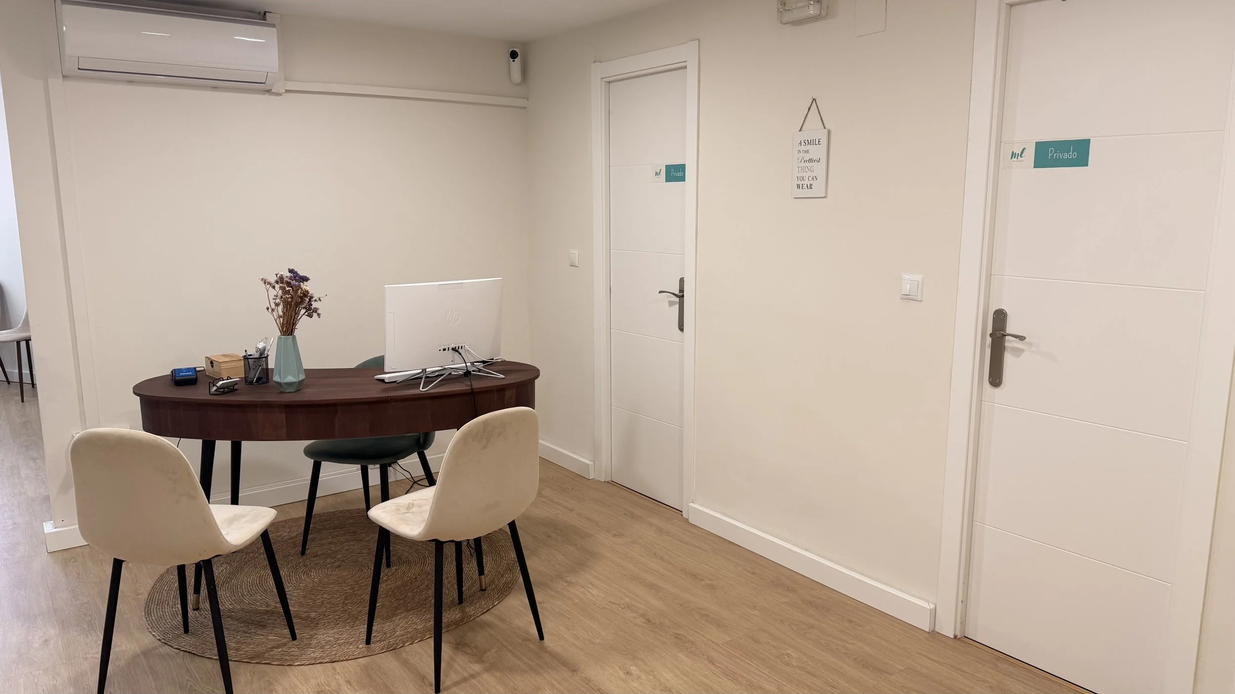 Sala de espera o oficina con mesa de madera, cuatro sillas de tela beige, un florero con flores y algunos objetos en la mesa, paredes blancas con letreros que dicen ‘Privado’ y ‘Me’ y una decoración en la pared.