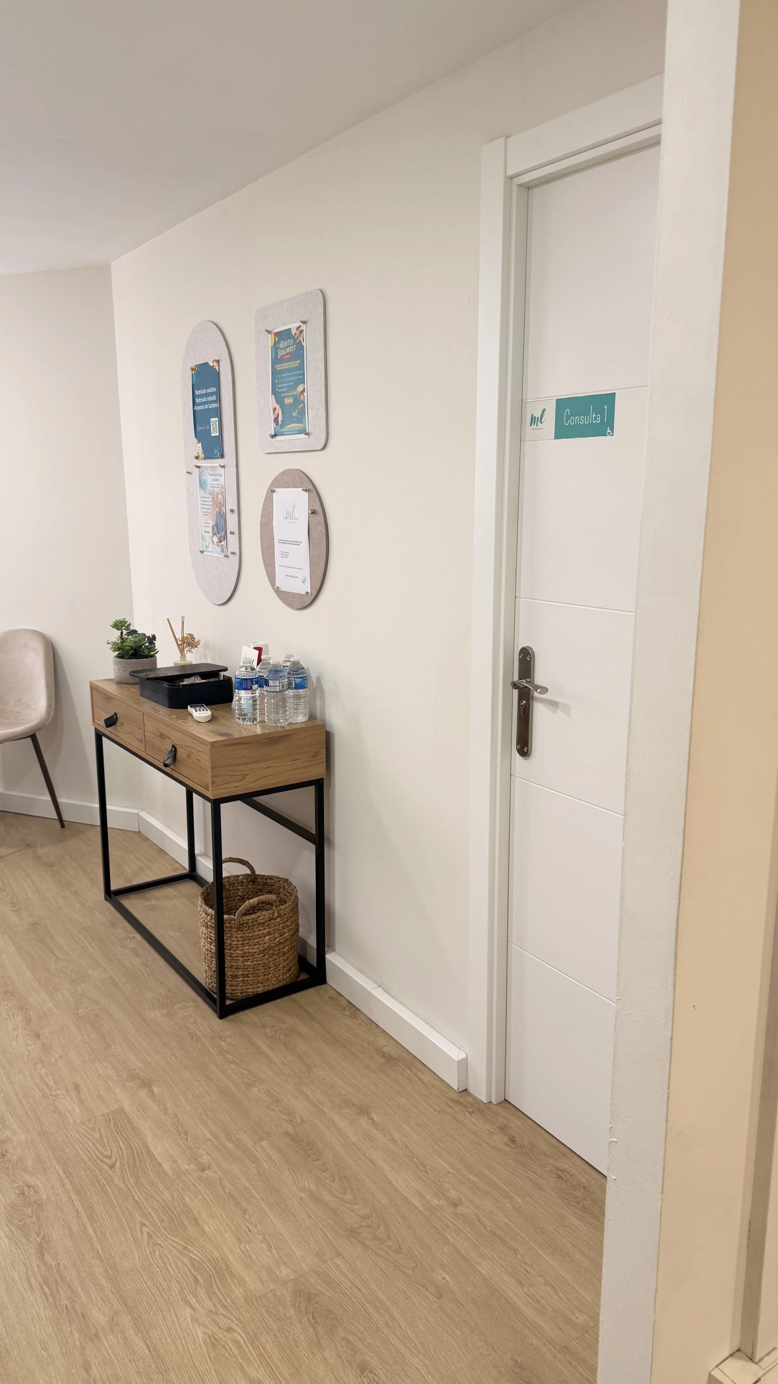 Sala de espera con pared blanca, cartelera con avisos en la pared, mesa con agua, plantas y otros objetos, cesta de mimbre en el suelo y puerta con señal que dice 'Consulta 1'.