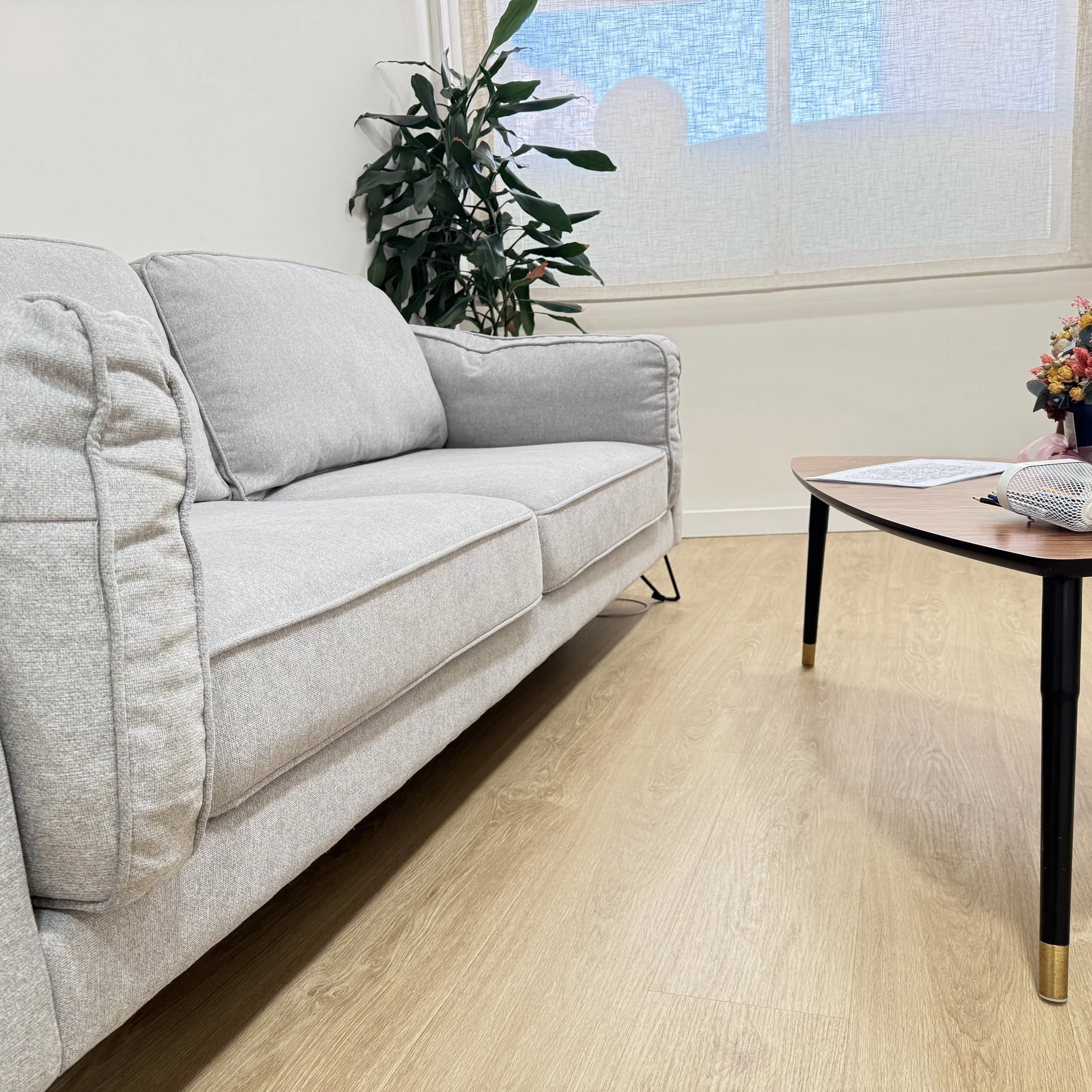 Sala de estar con sillón gris claro, planta verde en la esquina cerca de la ventana con cortina blanca, mesa de madera con libros y florero con flores coloridas.