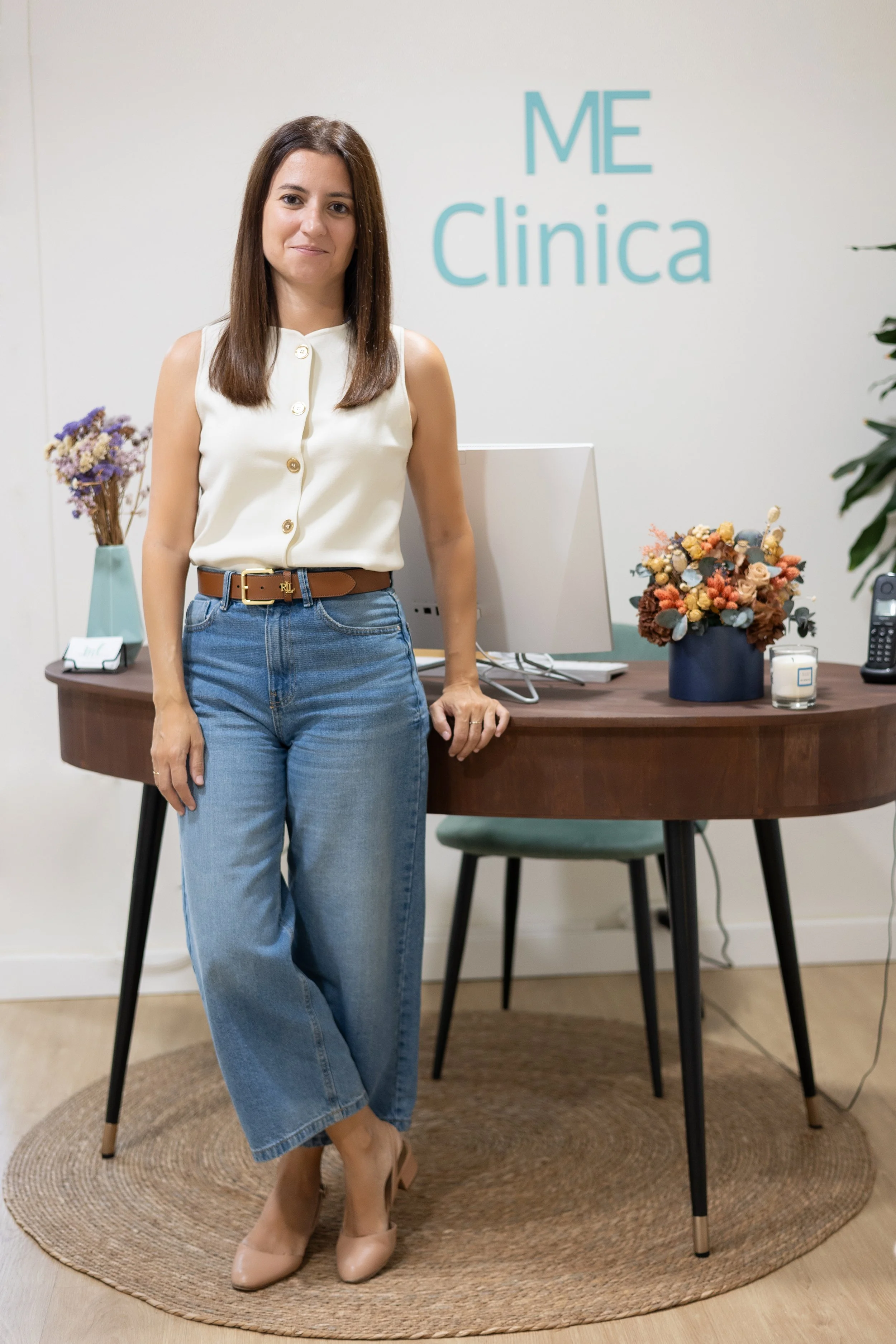 Mujer con blusa blanca, pantalones vaqueros y tacones beige, posando en una oficina moderna con letras que dicen "Me Clínica" en la pared trasera, decorada con flores y objetos de oficina.