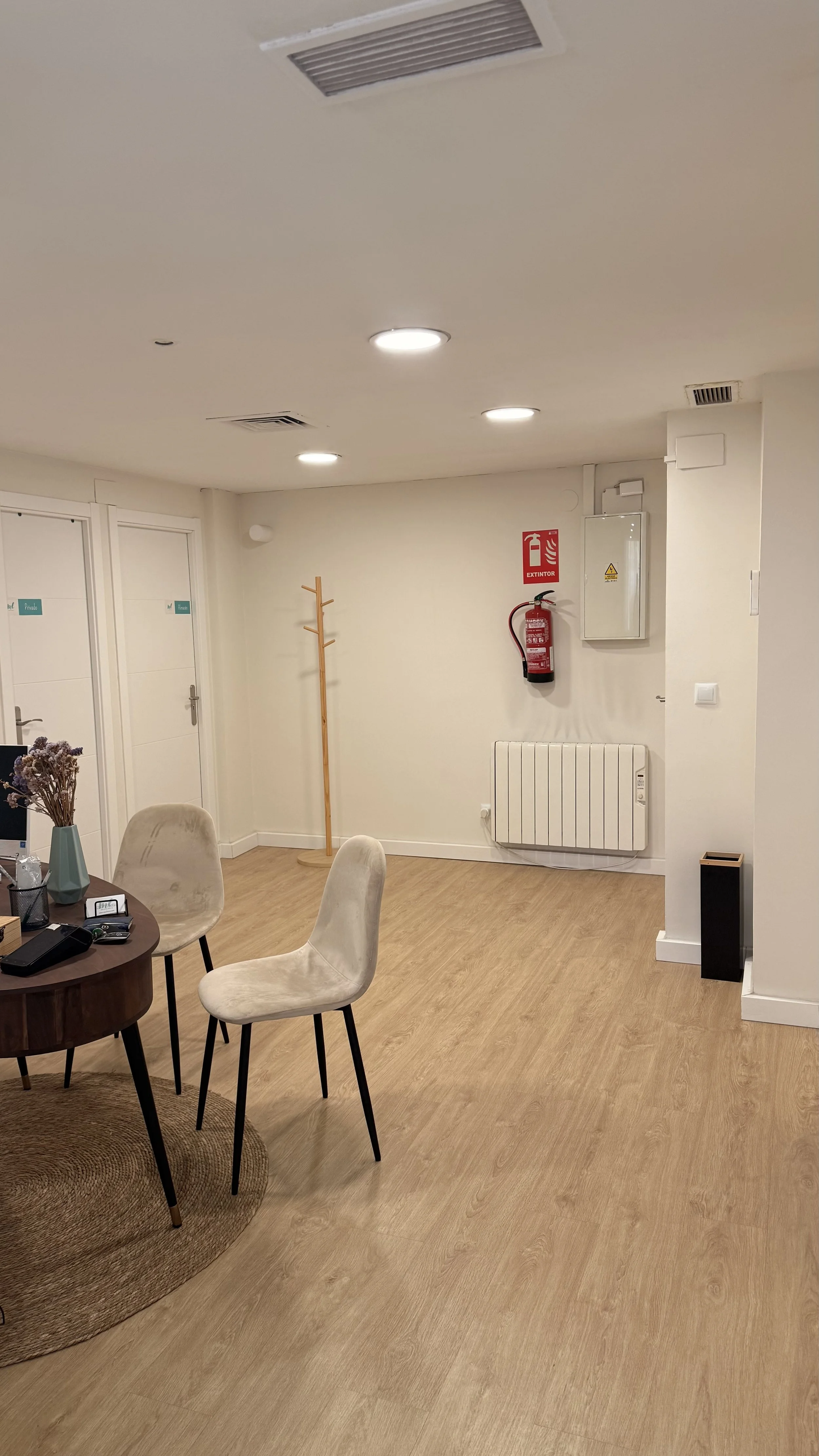 Una sala de oficina con una mesa redonda, sillas beige, una planta de laurel y en la pared un extintor, un radiador y un gabinete de seguridad contra incendios