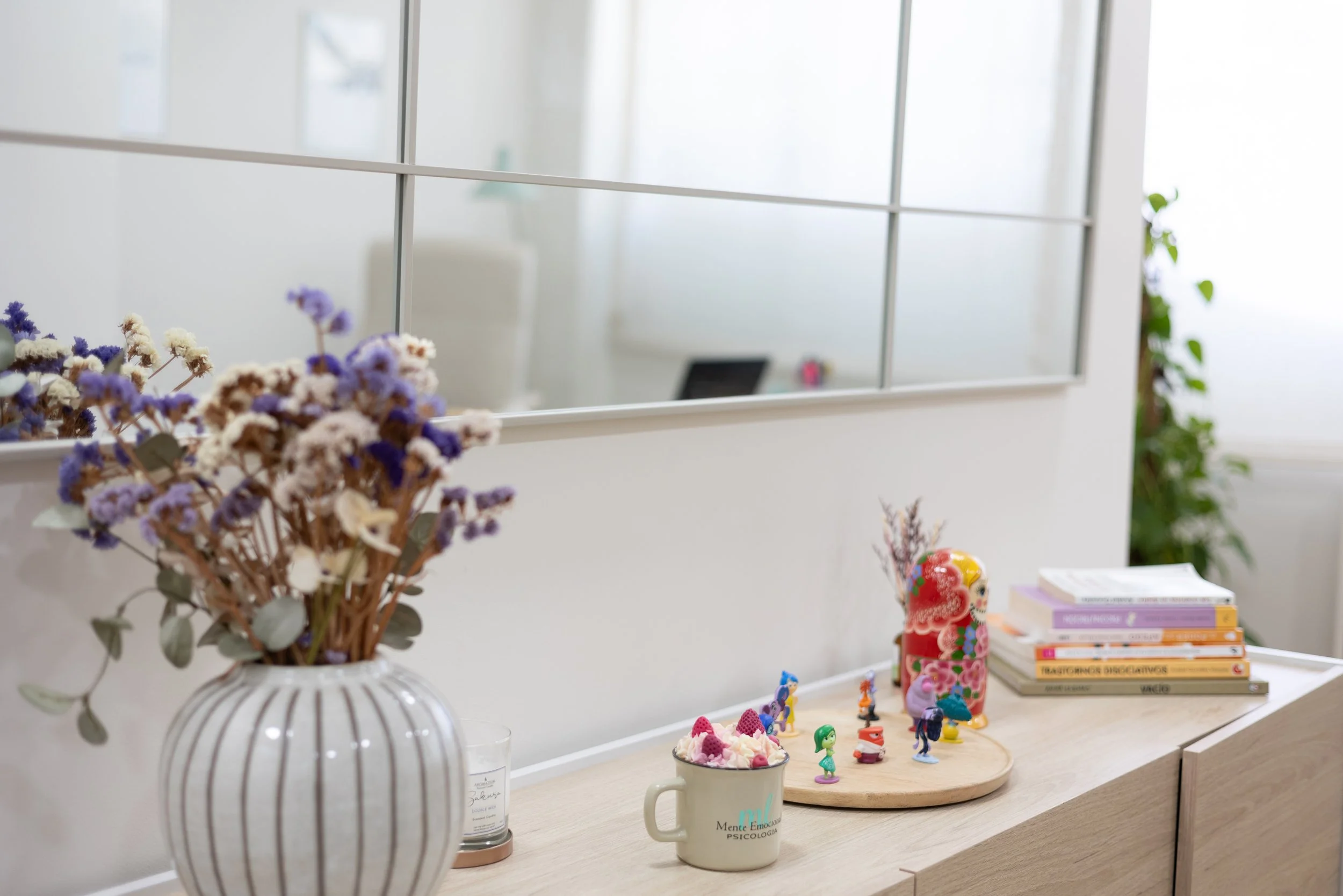 Vista de un escritorio con un florero con flores secas en primer plano, y en segundo plano hay figuras de plástico coloridas, una pila de libros y una taza. Detrás, un espejo en una pared blanca y algunas plantas verdes.