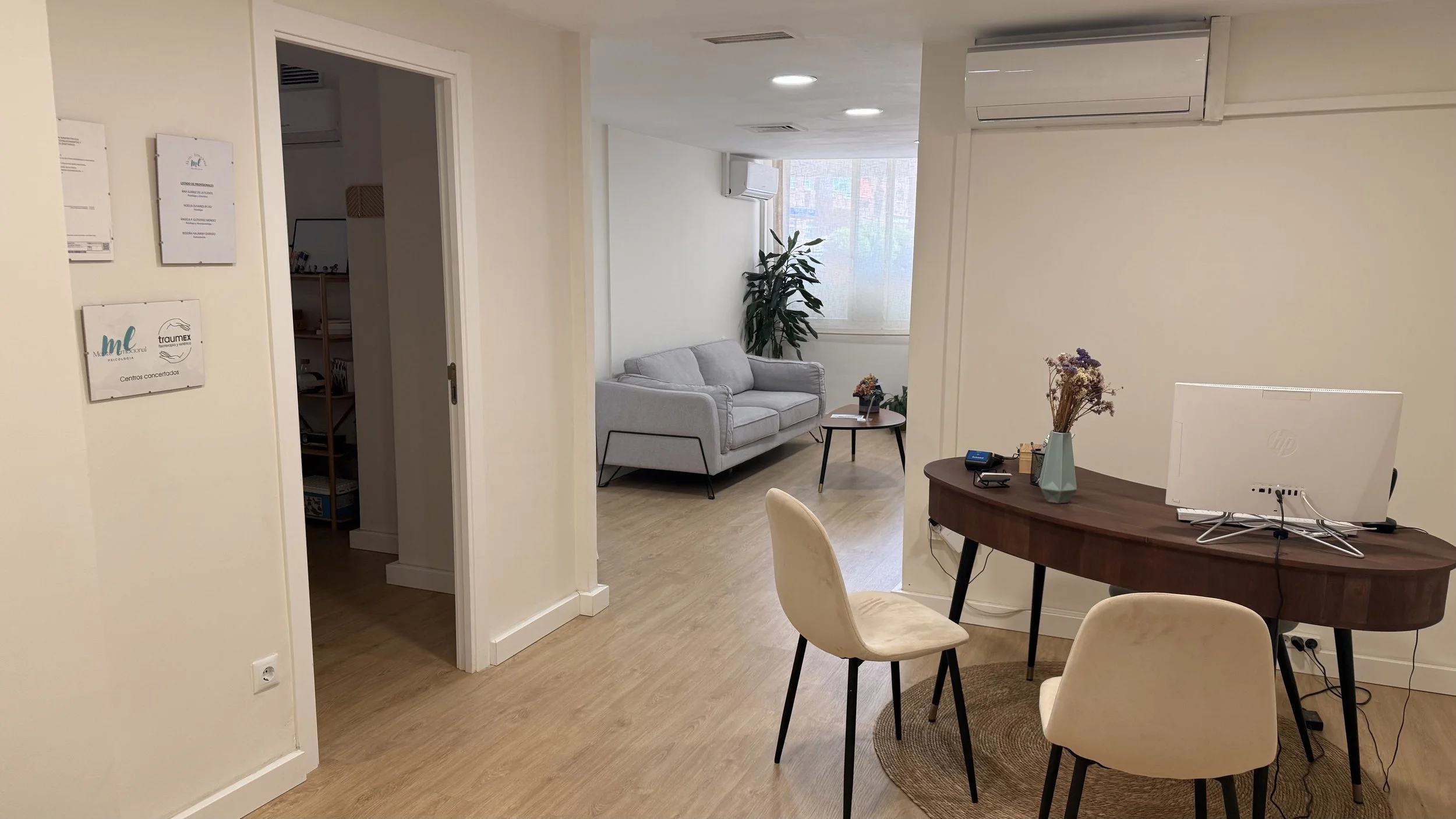 Espacio de oficina con zona de recepción, mesa con una computadora, sillas y una sala de espera con sofá y plantas, iluminación artificial y natural