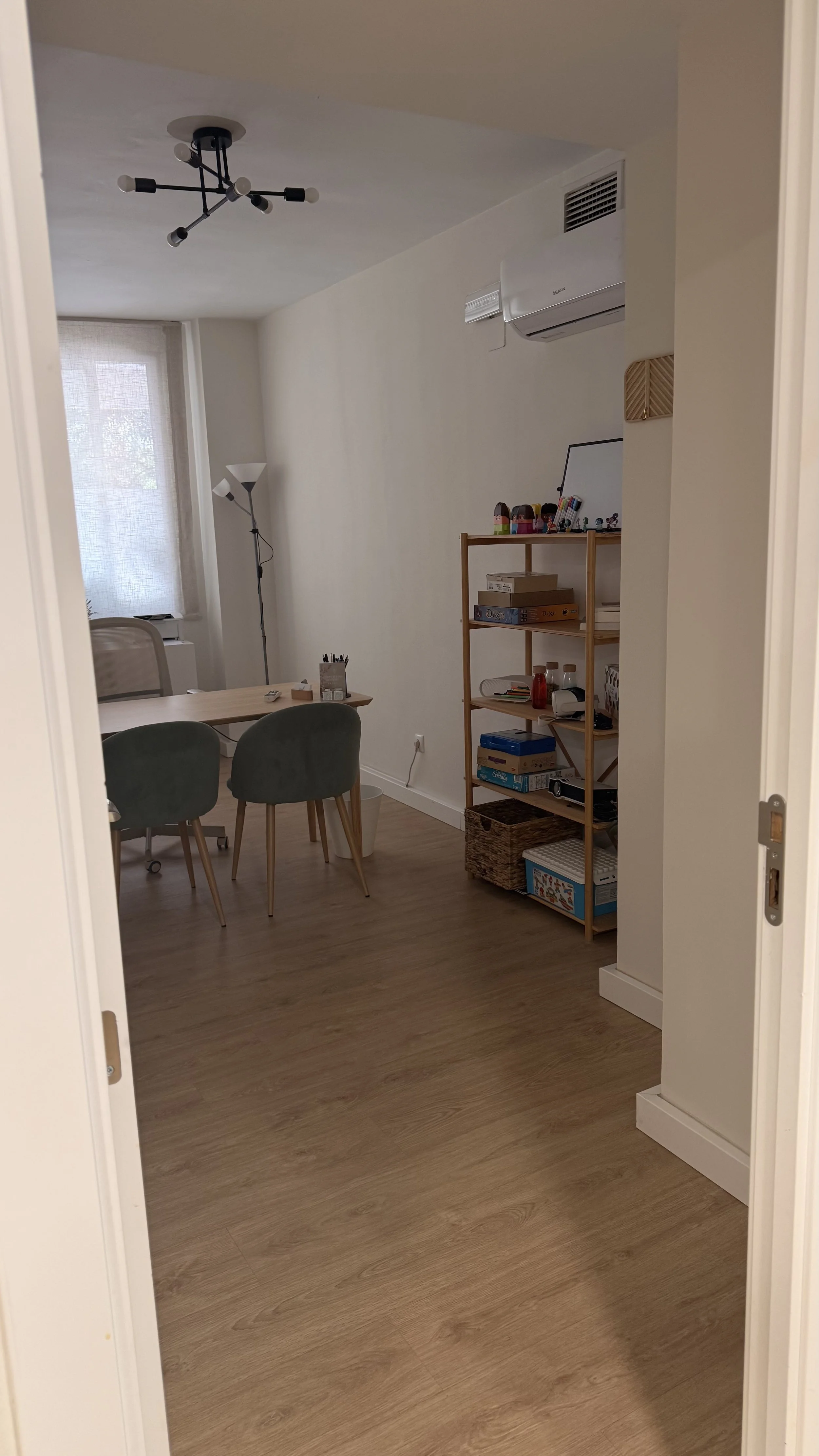 Vista de una sala de estar moderna con mesa de comedor, sillas verdes, estantería con libros y objetos, y una ventana con cortinas, con piso de madera y paredes blancas.