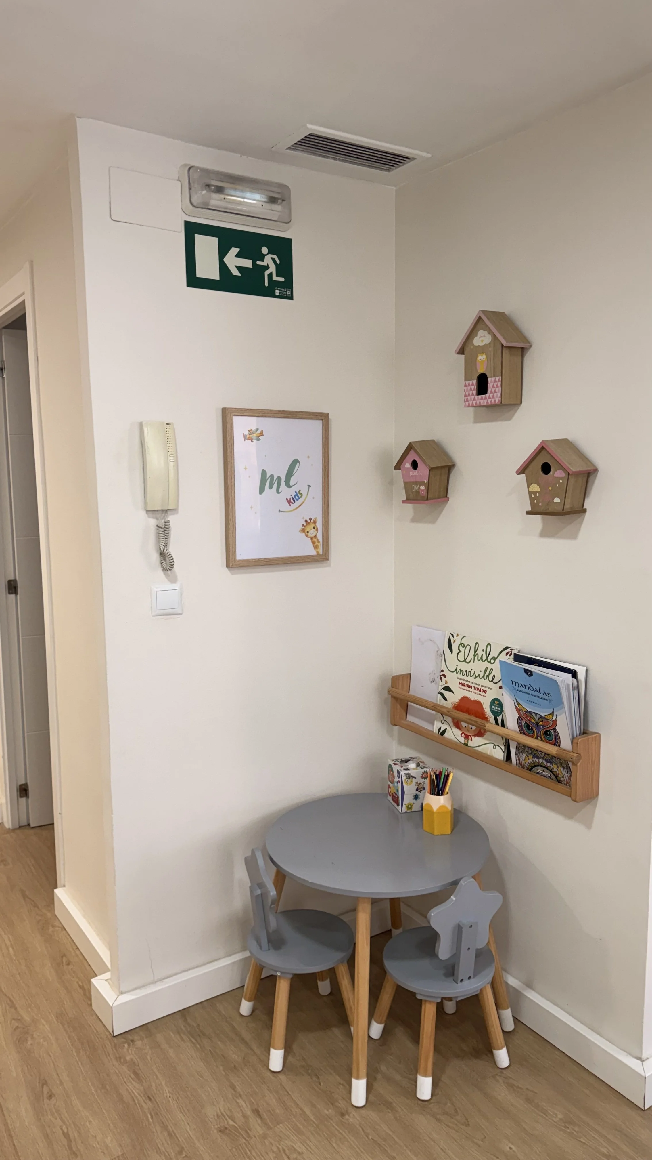 Rincón infantil con mesas y sillas de madera gris, estantería con libros, cuadros infantiles y aves de madera decorativas en la pared, señal de salida de emergencia y teléfono en la pared.