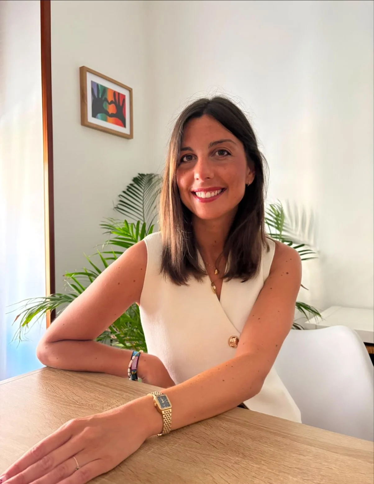 Una mujer sonriendo, con cabello castaño oscuro, usando vestido beige y accesorios dorados, en un ambiente con plantas verdes y un cuadro colorido en la pared.