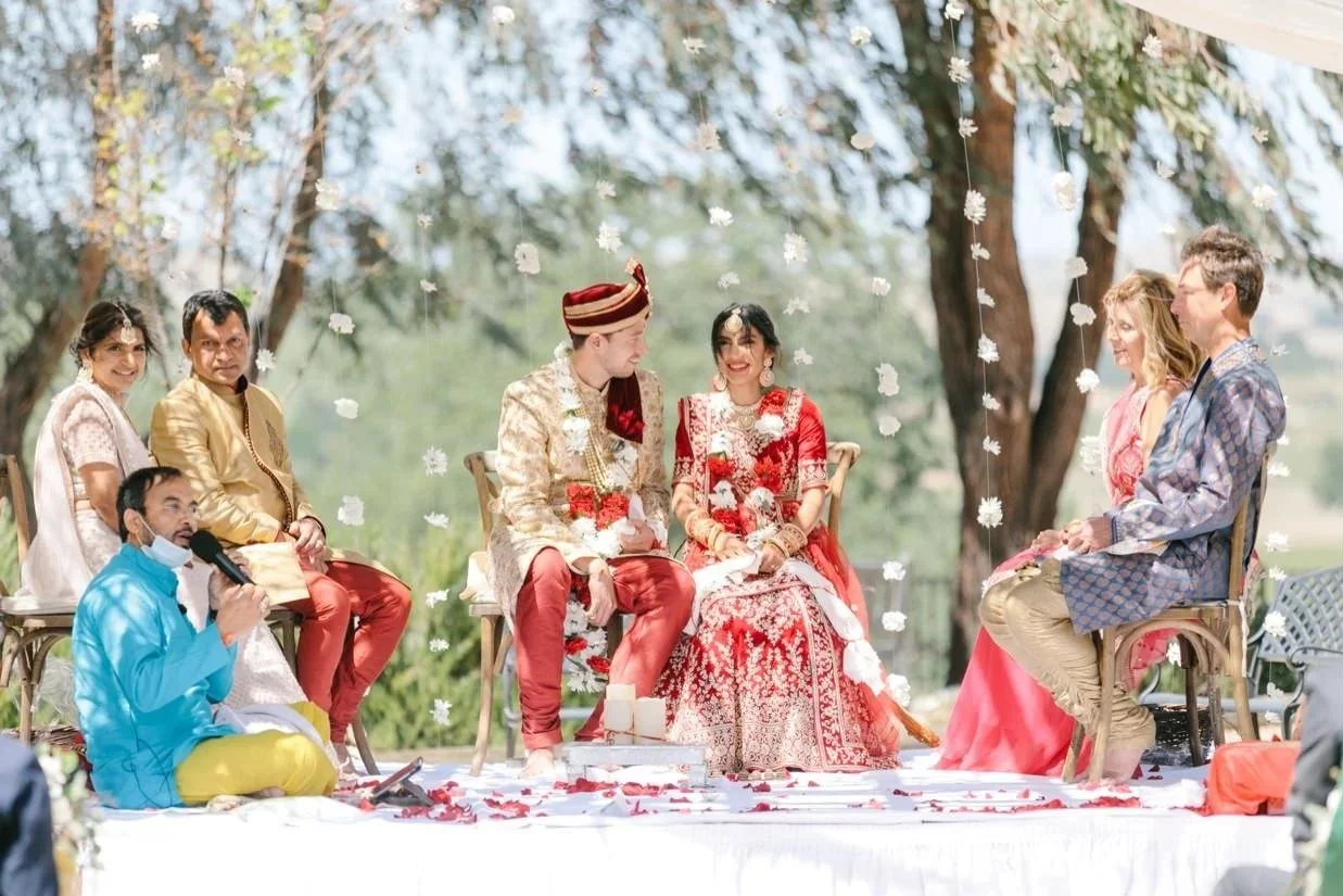 A traditional outdoor Indian wedding ceremony with a bride and groom seated in front of friends and family, surrounded by hanging flower decorations and petals on the ground at Four Sisters Ranch in Paso Robles, California.