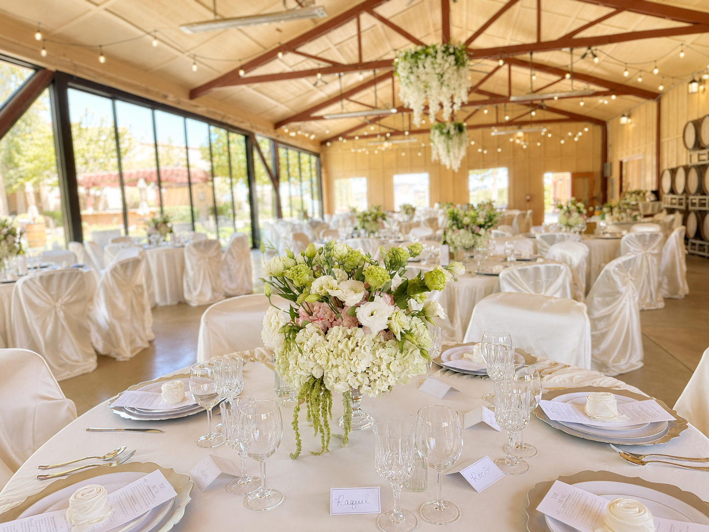 Elegant event space with round tables covered in white tablecloths, decorated with floral centerpieces, set with glassware, plates, and utensils. Chairs are draped in white covers, and a large floral chandelier hangs from the ceiling. The venue has l