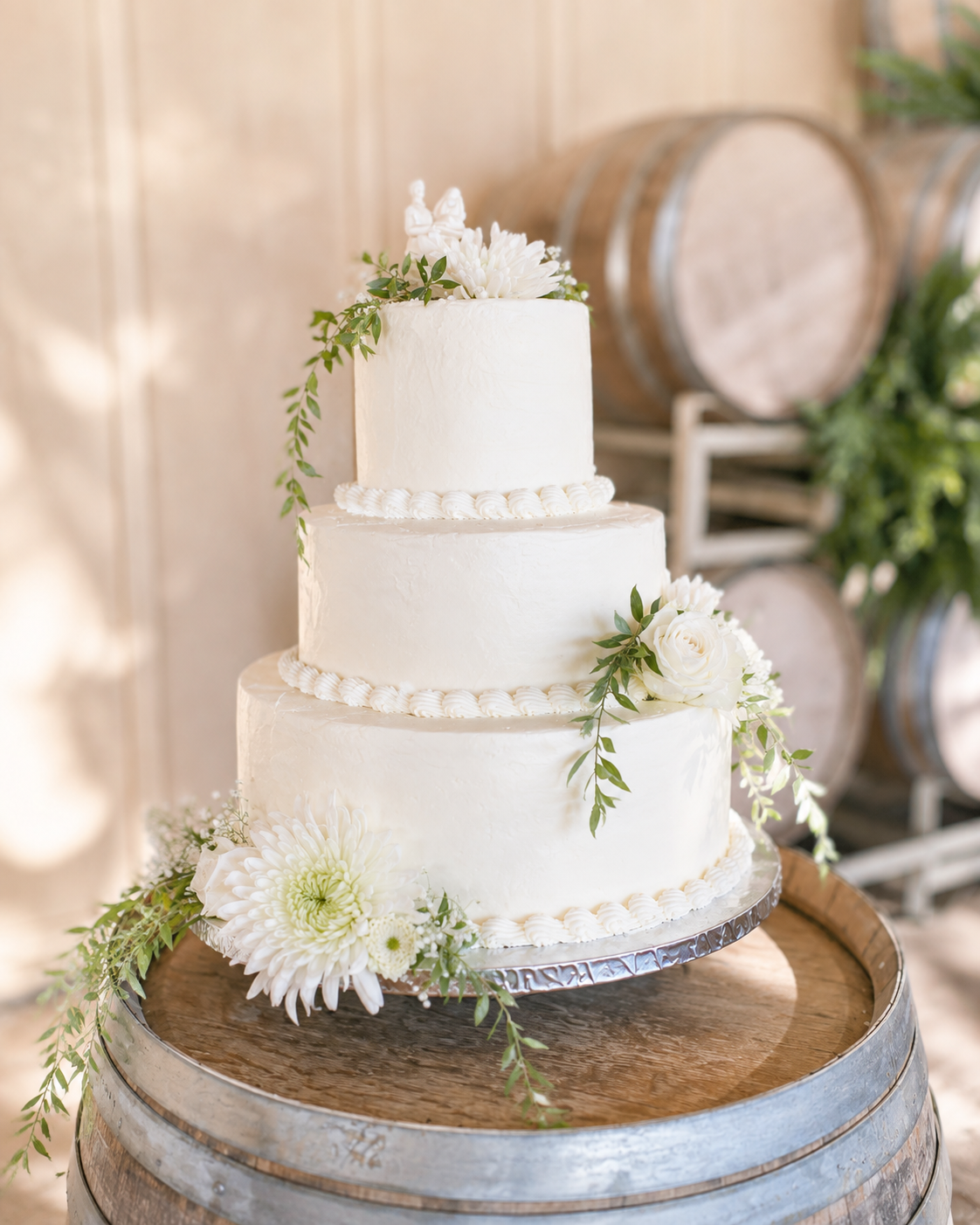 Three-tier white wedding cake decorated with white flowers and greenery, placed on a wooden barrel at Four Sisters Ranch wedding venue in wine country, California.