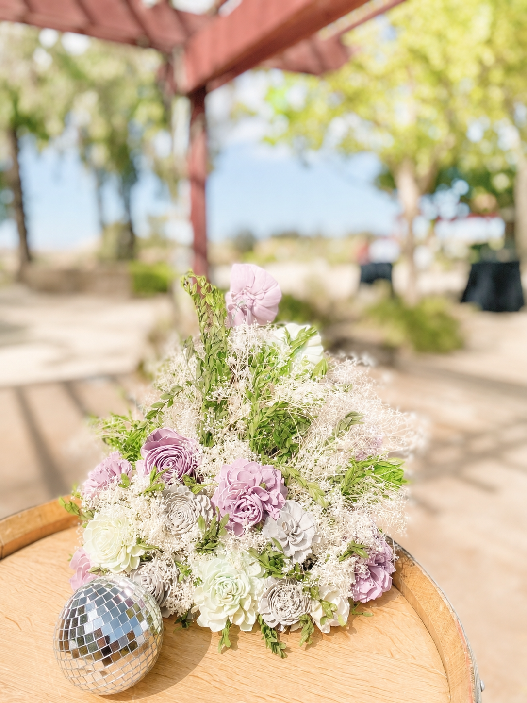 A bouquet of pastel-colored flowers and a disco ball on a wooden surface outdoors with trees at Four Sisters Ranch wedding venue.