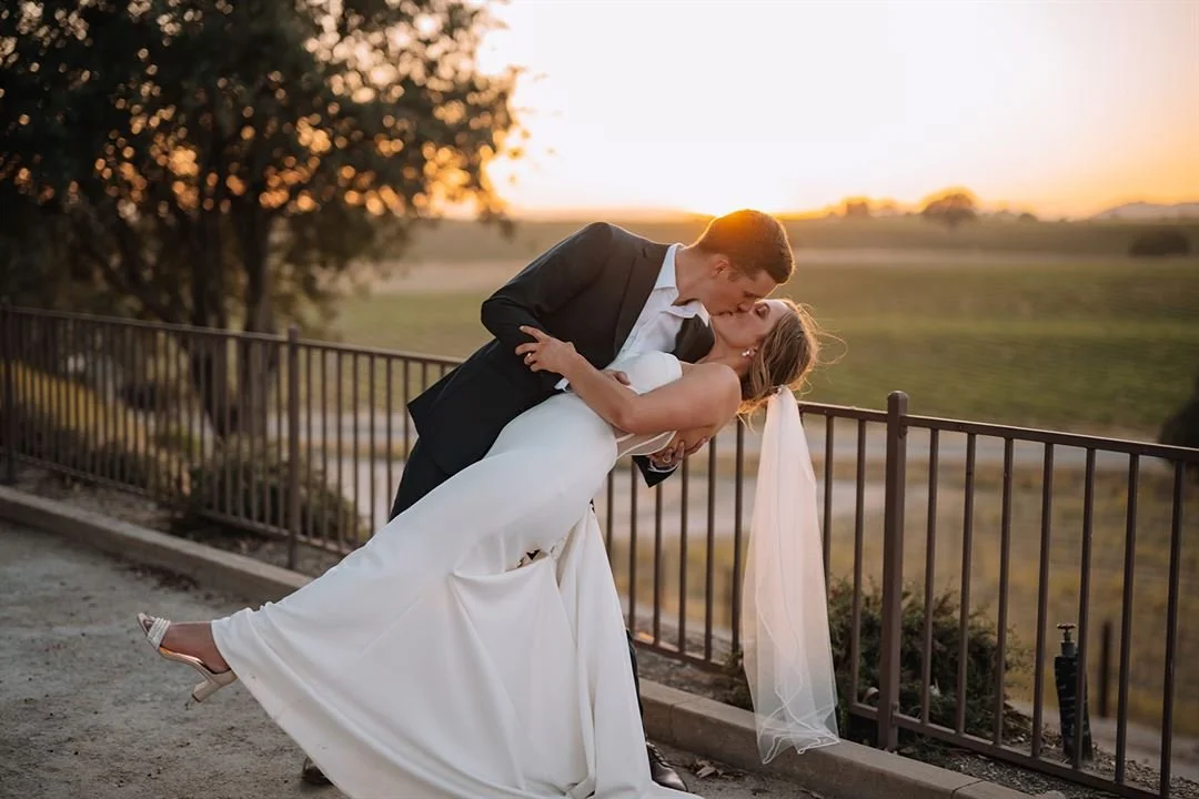 A newlywed couple sharing a kiss outdoors at sunset, with the groom dipping the bride in front of a fence and scenic countryside in the background.