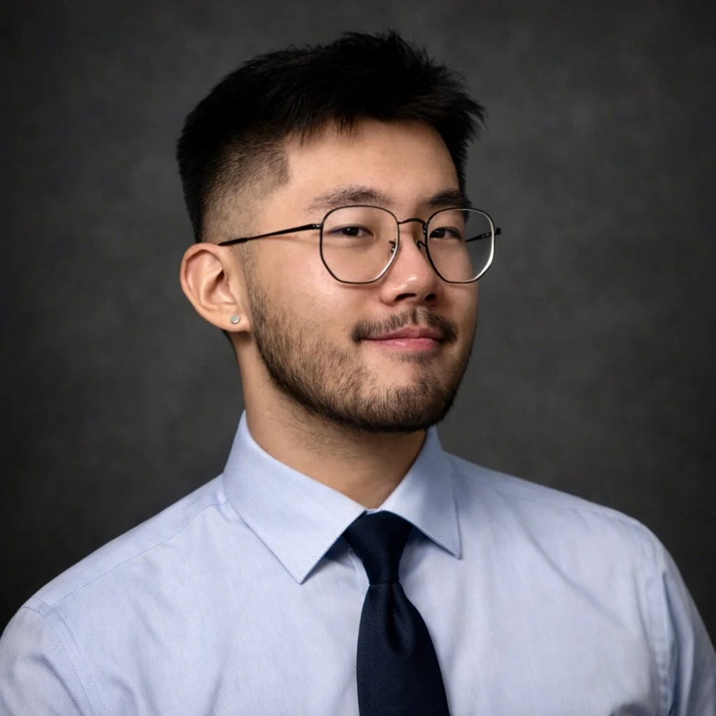 A young man with glasses, a beard, and short dark hair, wearing a light blue dress shirt and a dark blue tie, poses for a professional headshot against a dark backdrop.