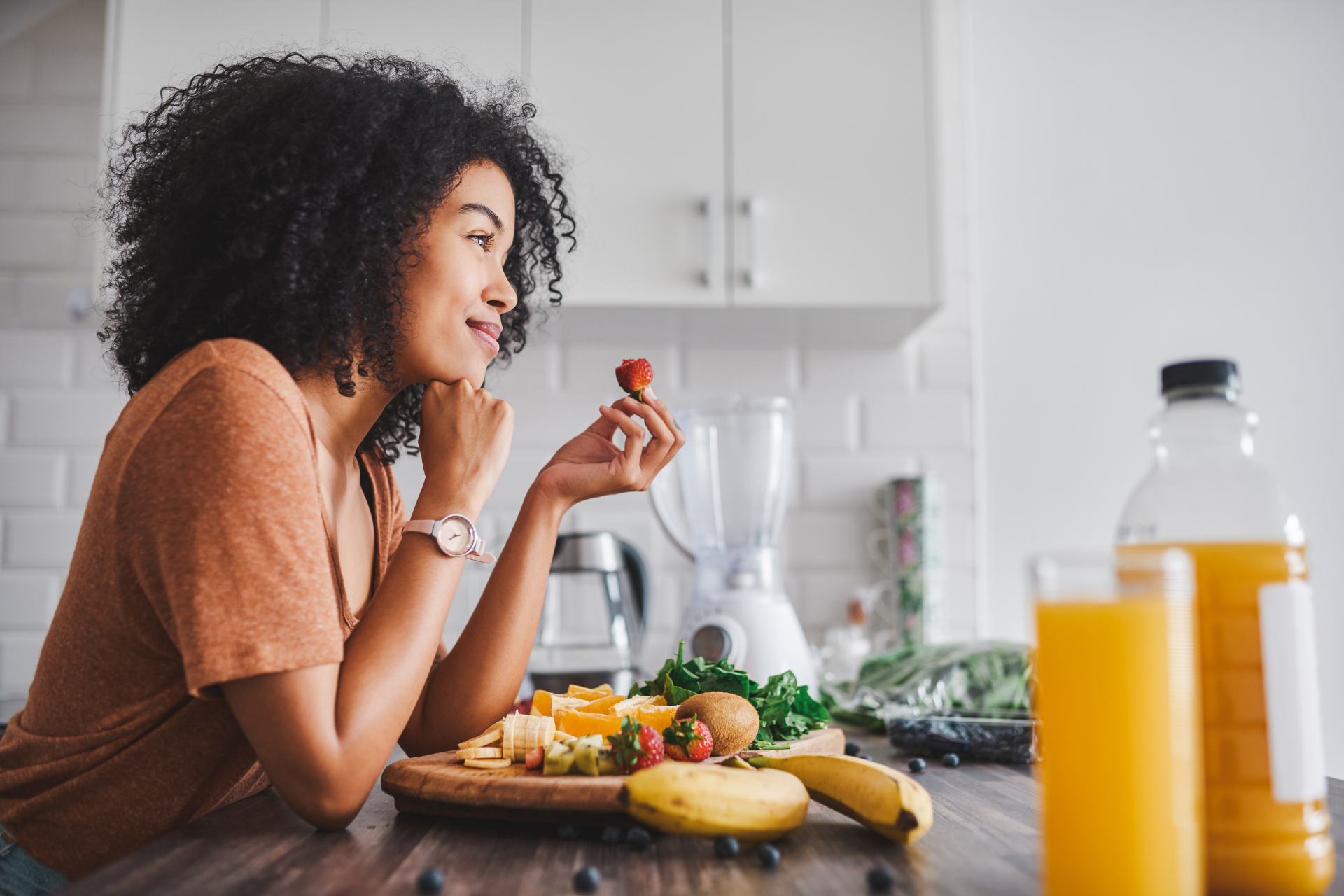 A woman with curly hair in a brown top holds a strawberry while sitting at a kitchen counter with sliced fruit, greens, and baked goods on a wooden cutting board, with orange juice bottles nearby.