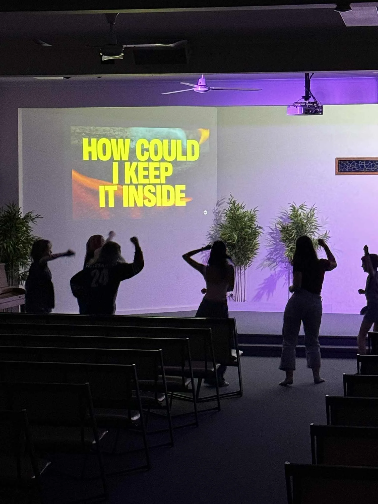 Music equipment including a keyboard, drums, microphone, and amplifiers set up in front of a white wall decorated with a green cross and plants, suggesting a church or worship space.
