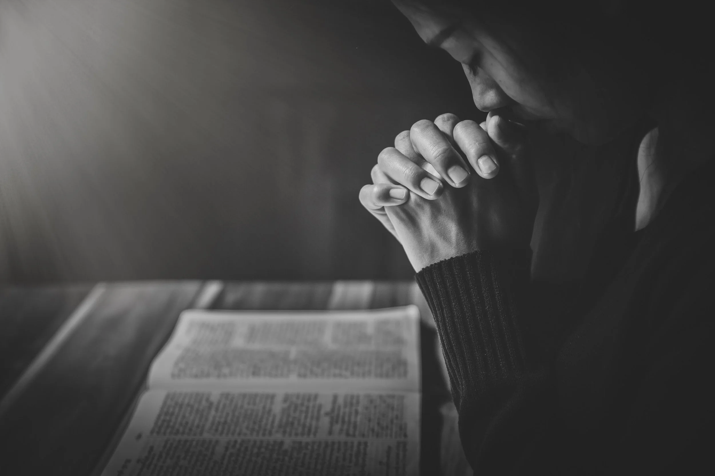 A person in prayer with hands clasped, head bowed, sitting at a table with an open book, in black and white.