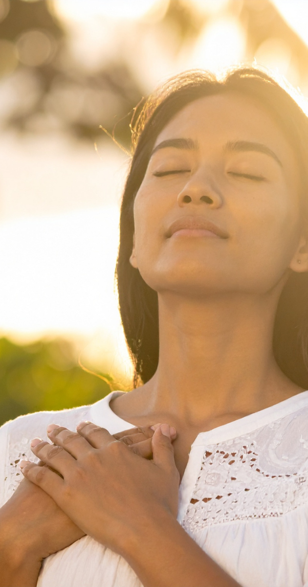 A woman with medium length dark hair wearing a white long sleeve shirt is holding her hands to her chest in soft, outdoor lighting.