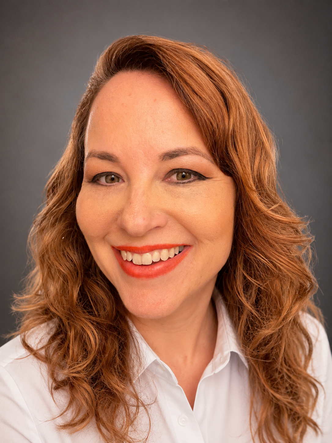 A woman with red curly hair smiling, wearing a white shirt, against a dark grey background.