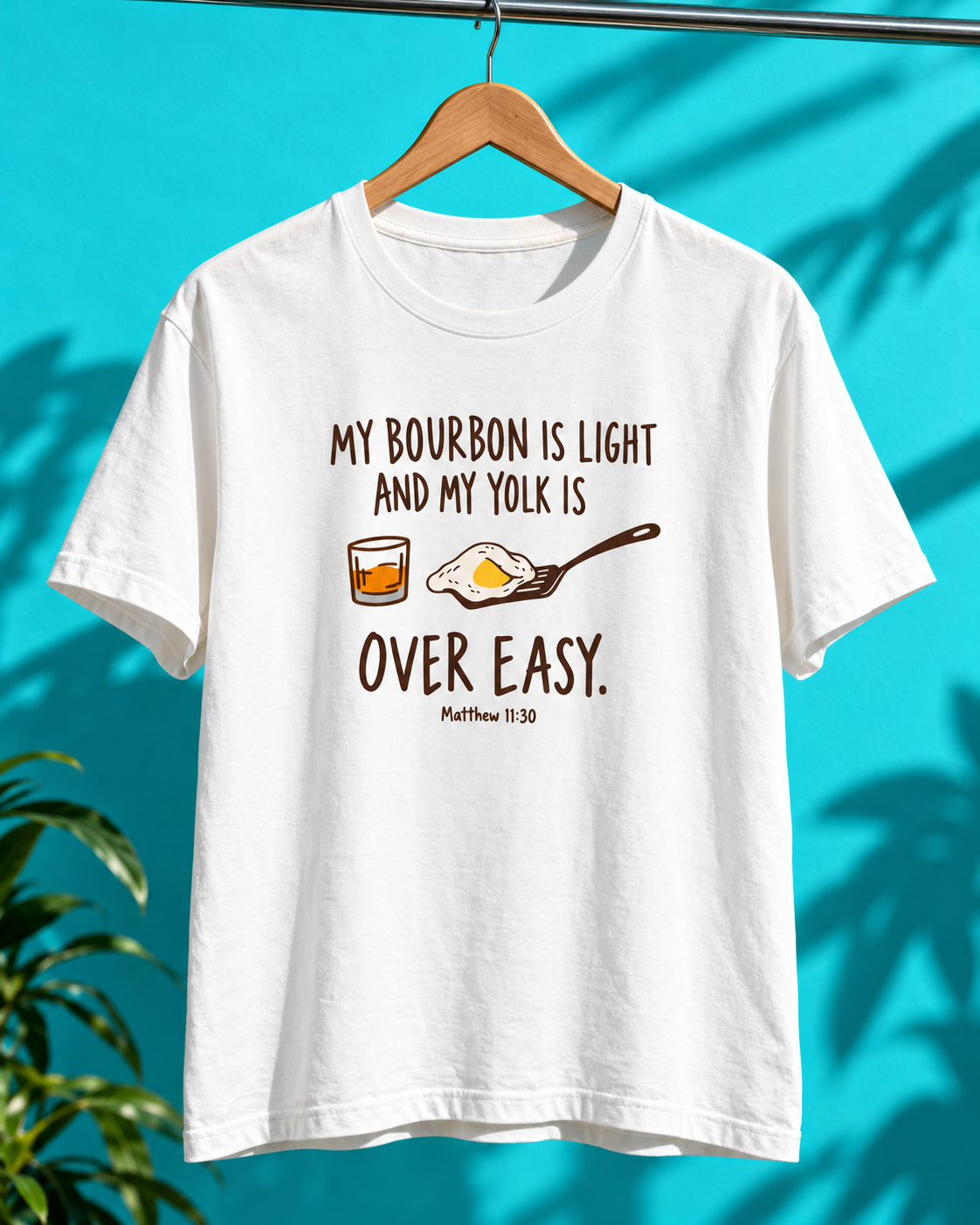 White T-shirt with bourbon glass and fried egg illustrations and the text “My bourbon is light and my yolk is over easy,” hanging against a turquoise background.