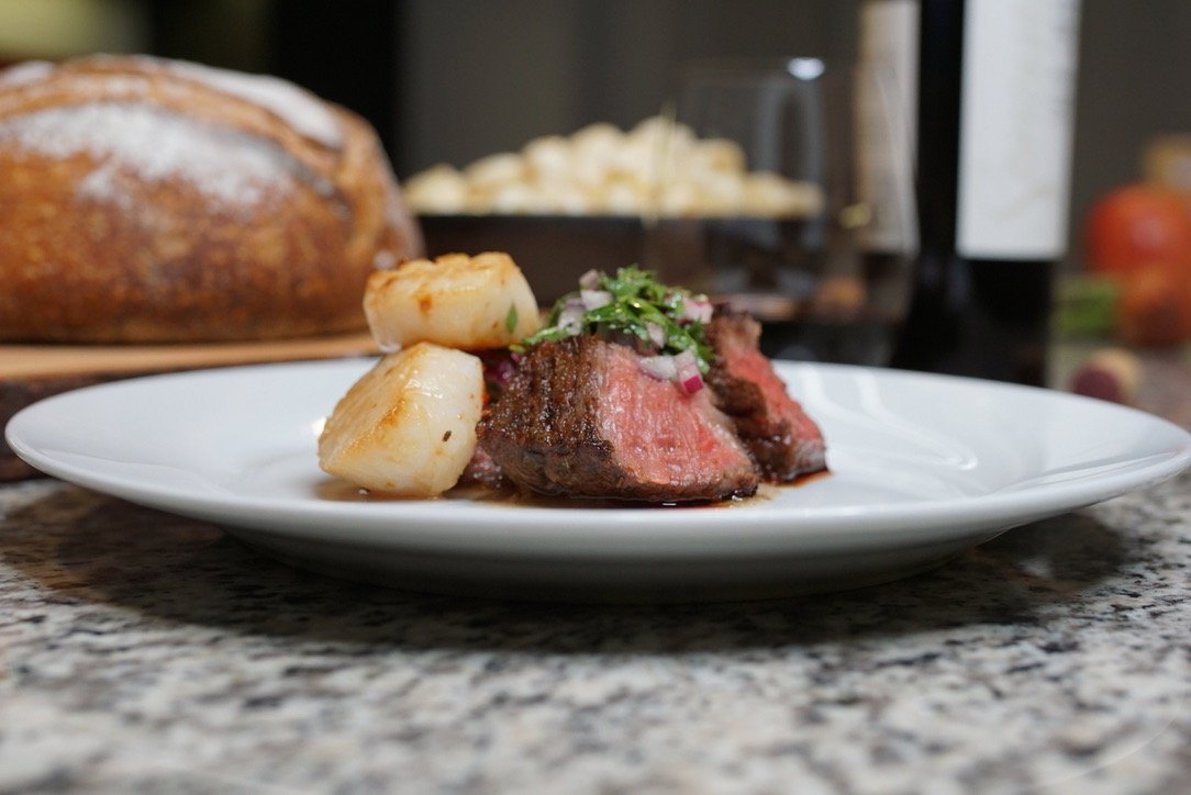 A white plate with sliced cooked steak topped with chopped green onions and red onions, alongside roasted scallops, on a kitchen countertop with bread and condiments in the background.
