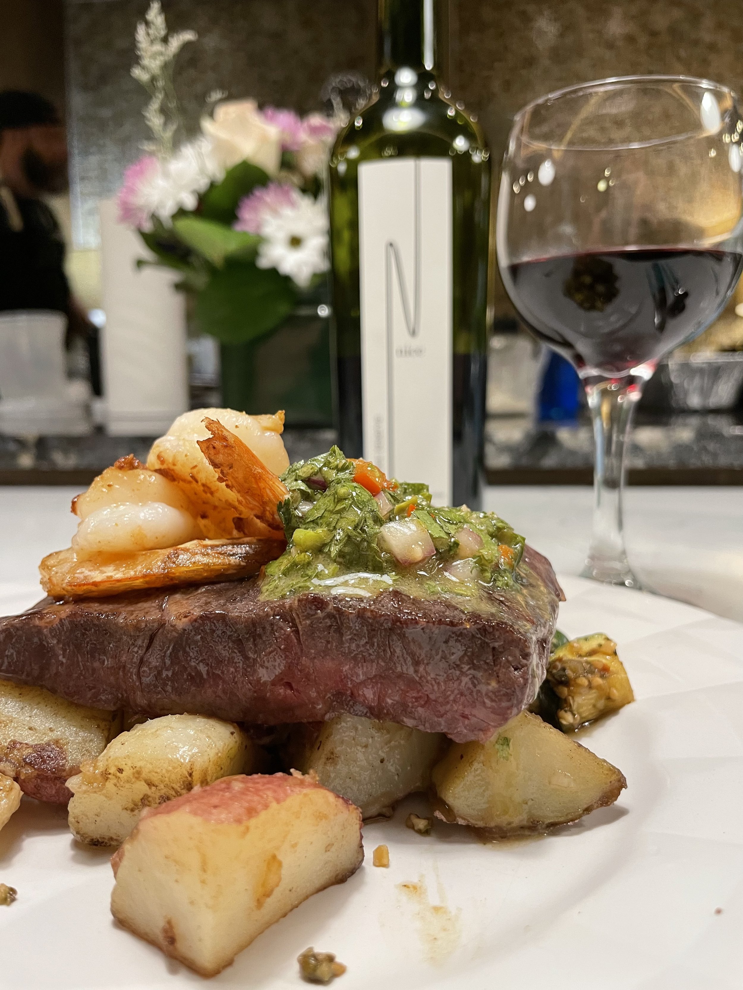 A plate of cooked steak topped with a green garnish and served with roasted potatoes. In the background, there is a glass of red wine, a bottle of wine, and a flower arrangement.