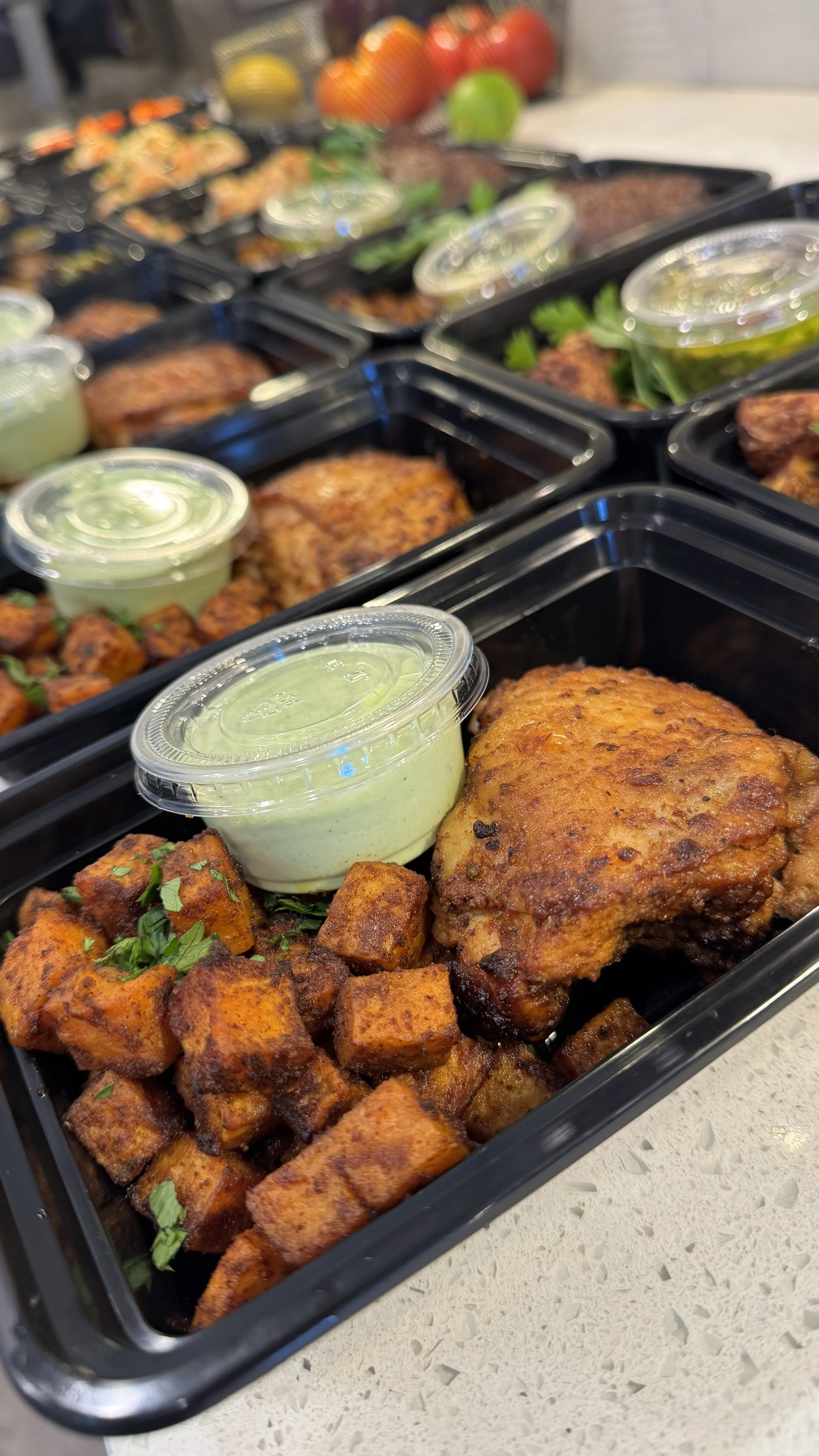 Black meal prep container with fried chicken thigh, roasted sweet potato cubes, and a small cup of green dipping sauce. In the background, additional containers with similar meals are displayed, along with fresh tomatoes, lemons, and limes.