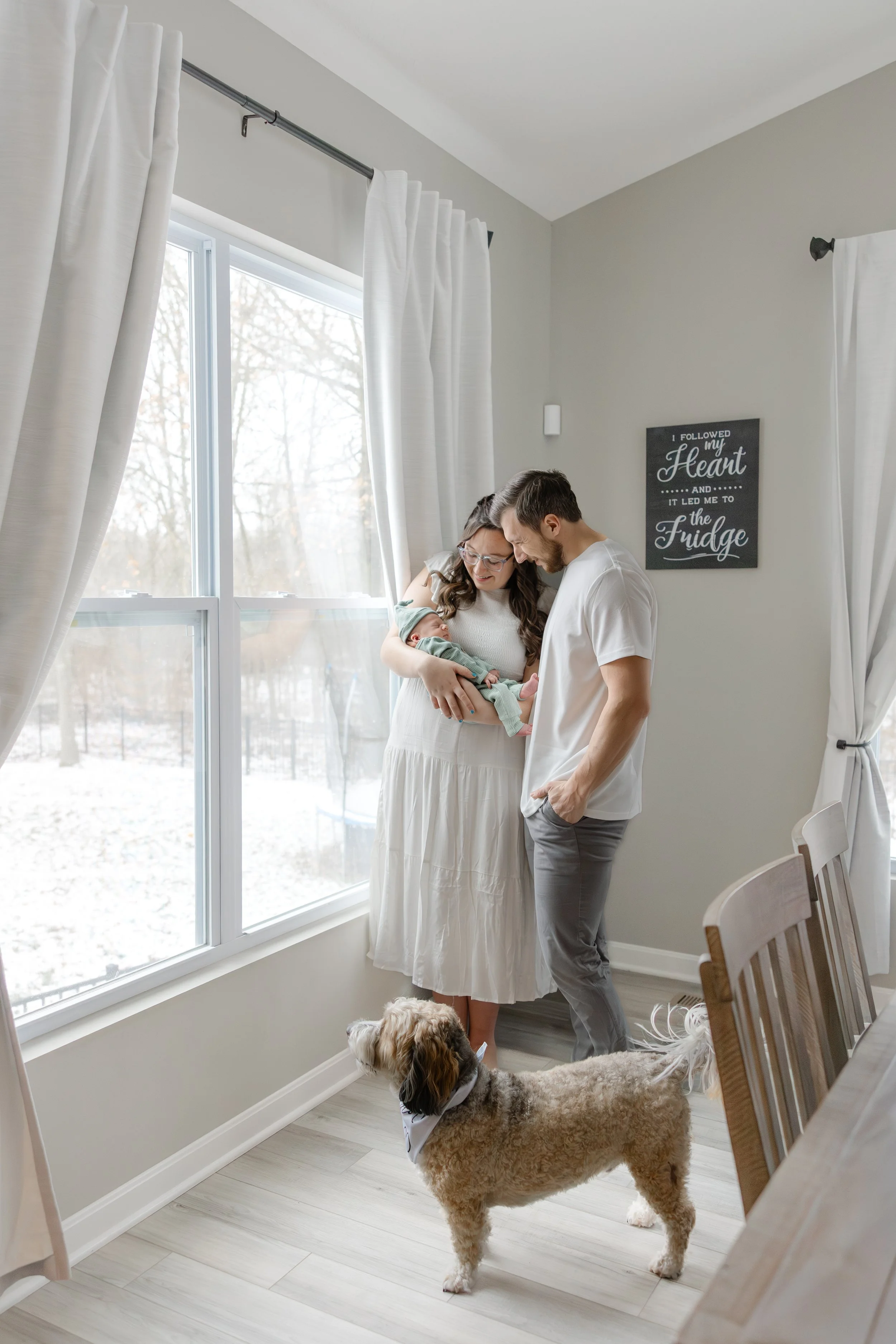A family of three, including a woman holding a newborn baby and a man, standing by a large window with white curtains, while their dog looks at them inside a bright room with light-colored walls and a sign on the wall reading 'I followed my heart and