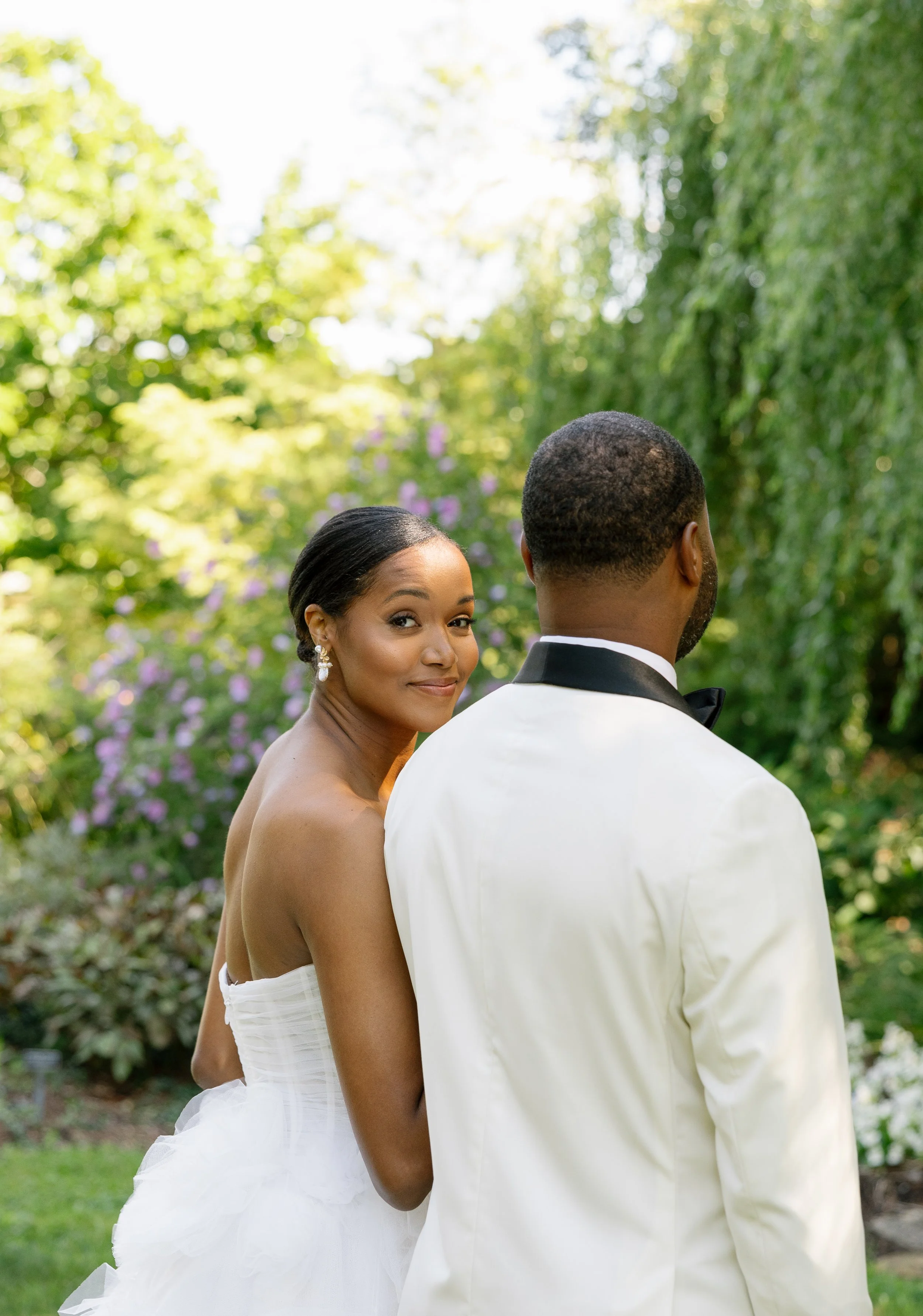 A bride and groom in wedding attire in a lush outdoor garden setting, with the bride smiling at the camera behind the groom turning away.