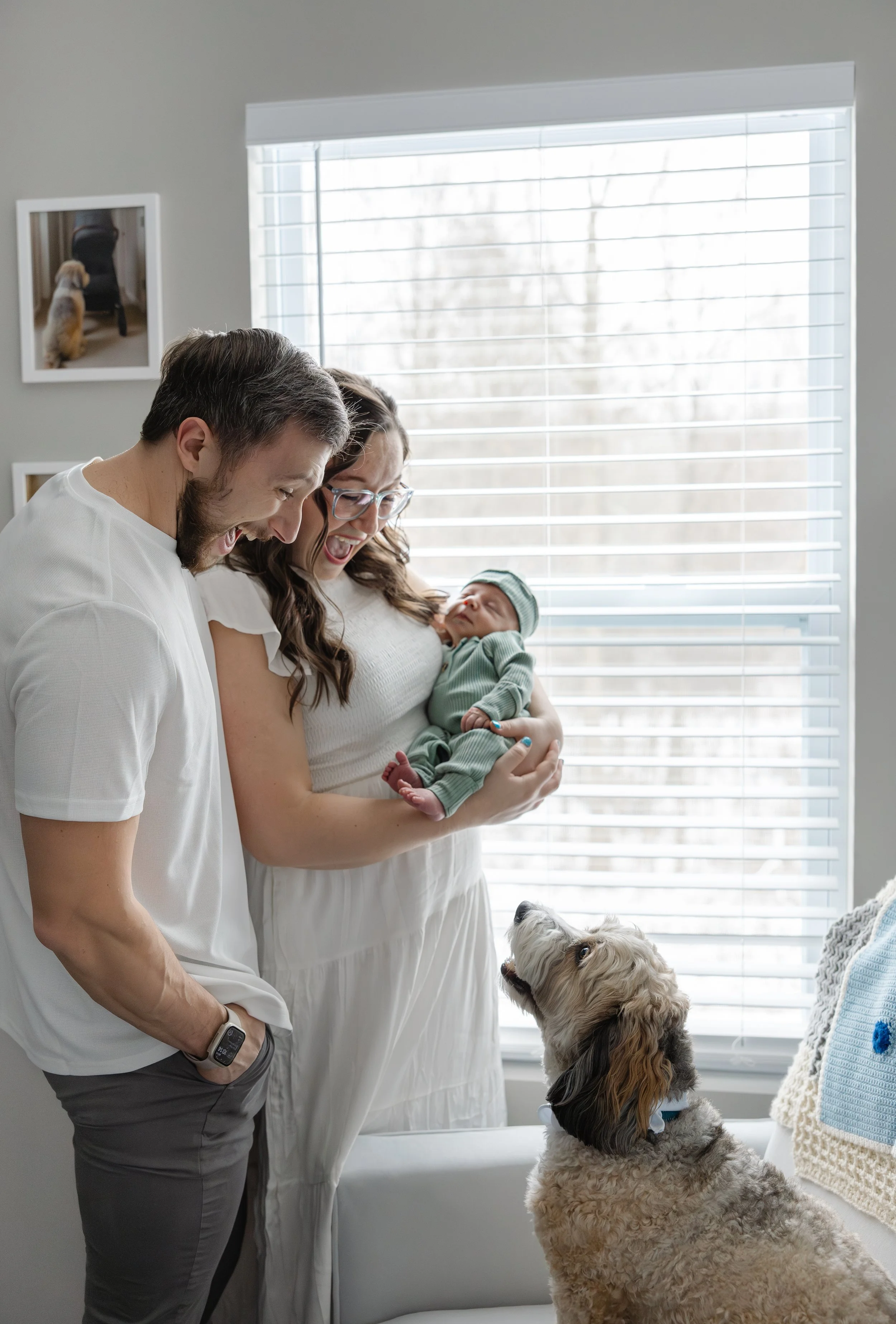 A happy family of three celebrating a new baby in a bright living room, with a dog looking up at them.