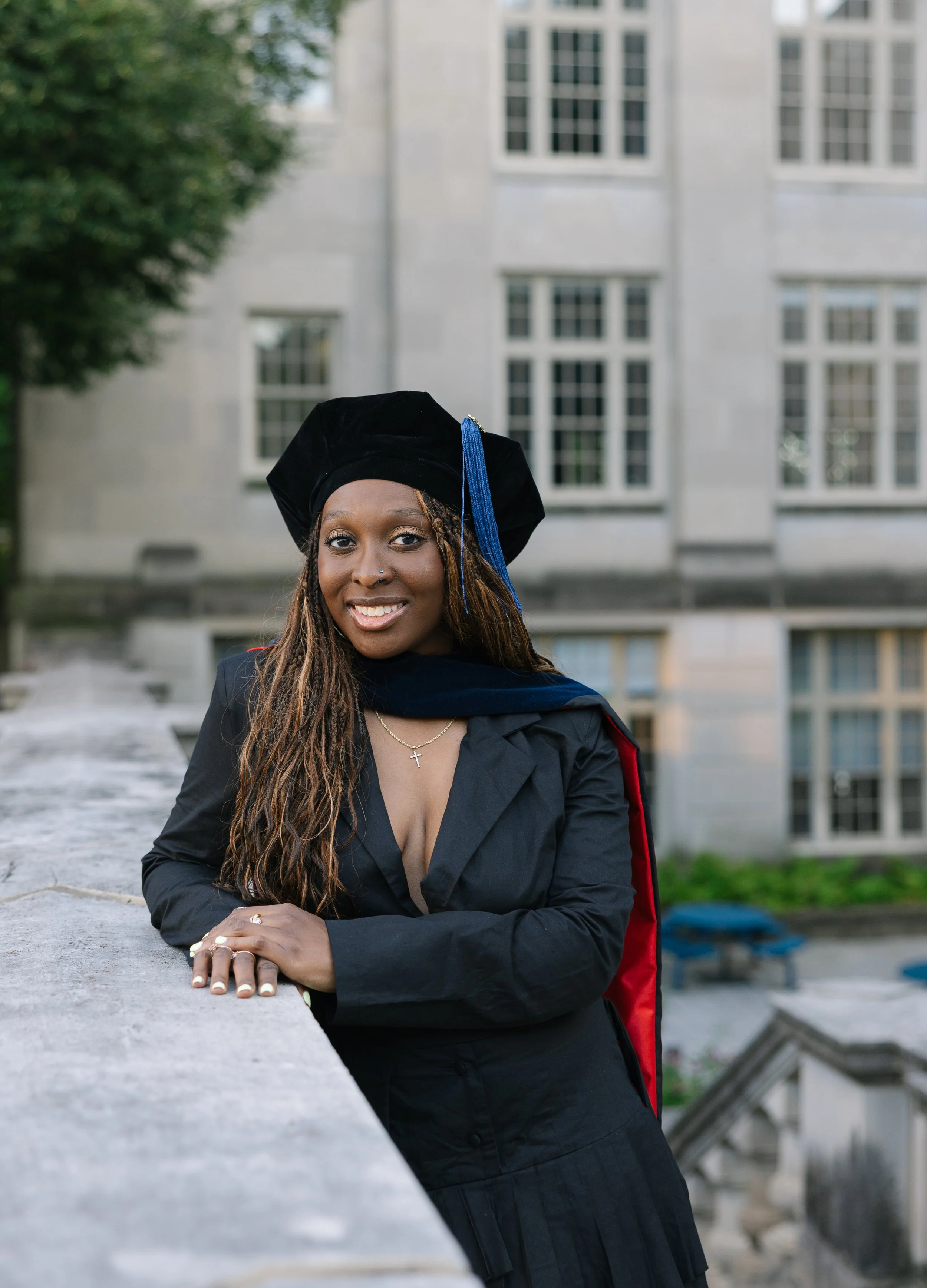 A woman in a graduation gown holding a cap, standing on the steps outside a building with large columns, smiling.