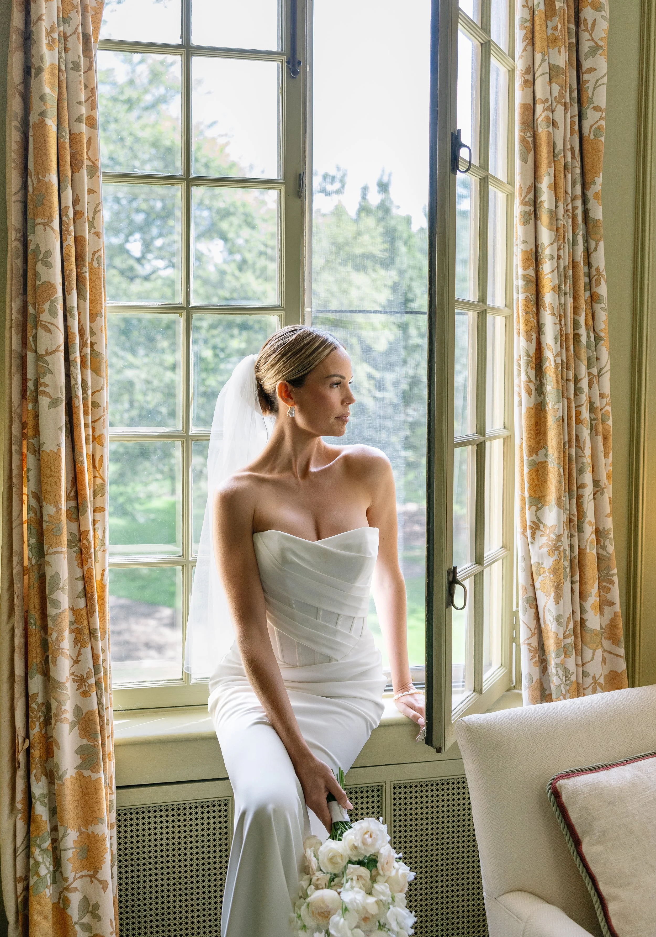 A bride in a strapless white wedding dress with a bouquet of white flowers sitting on a windowsill in a room with cream-colored floral curtains. She is looking thoughtfully out the window, with sunlight illuminating her profile and a view of greenery
