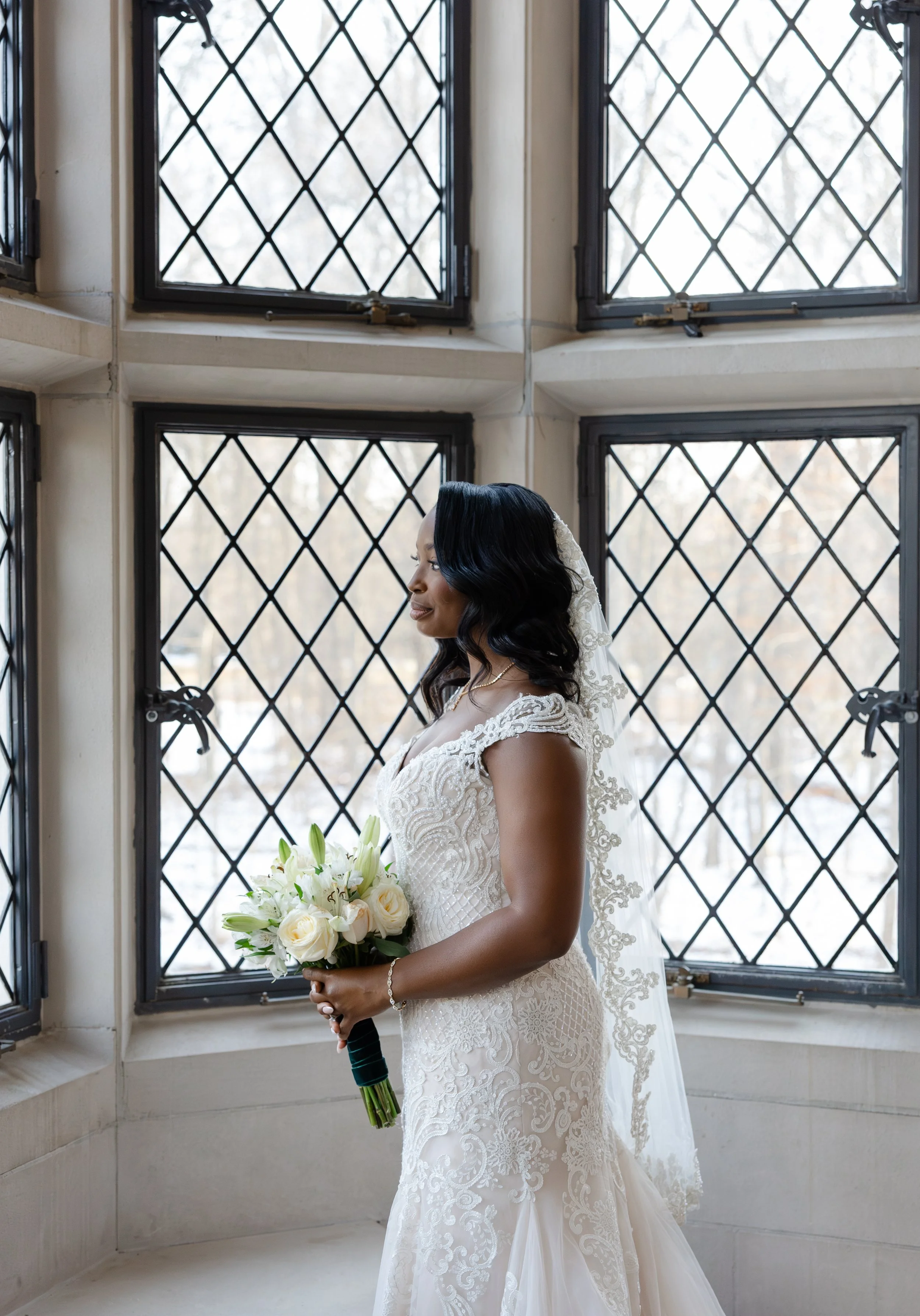 Bride in a lace wedding dress holding a bouquet of white roses and lilies, standing by a window with diamond-shaped panes.
