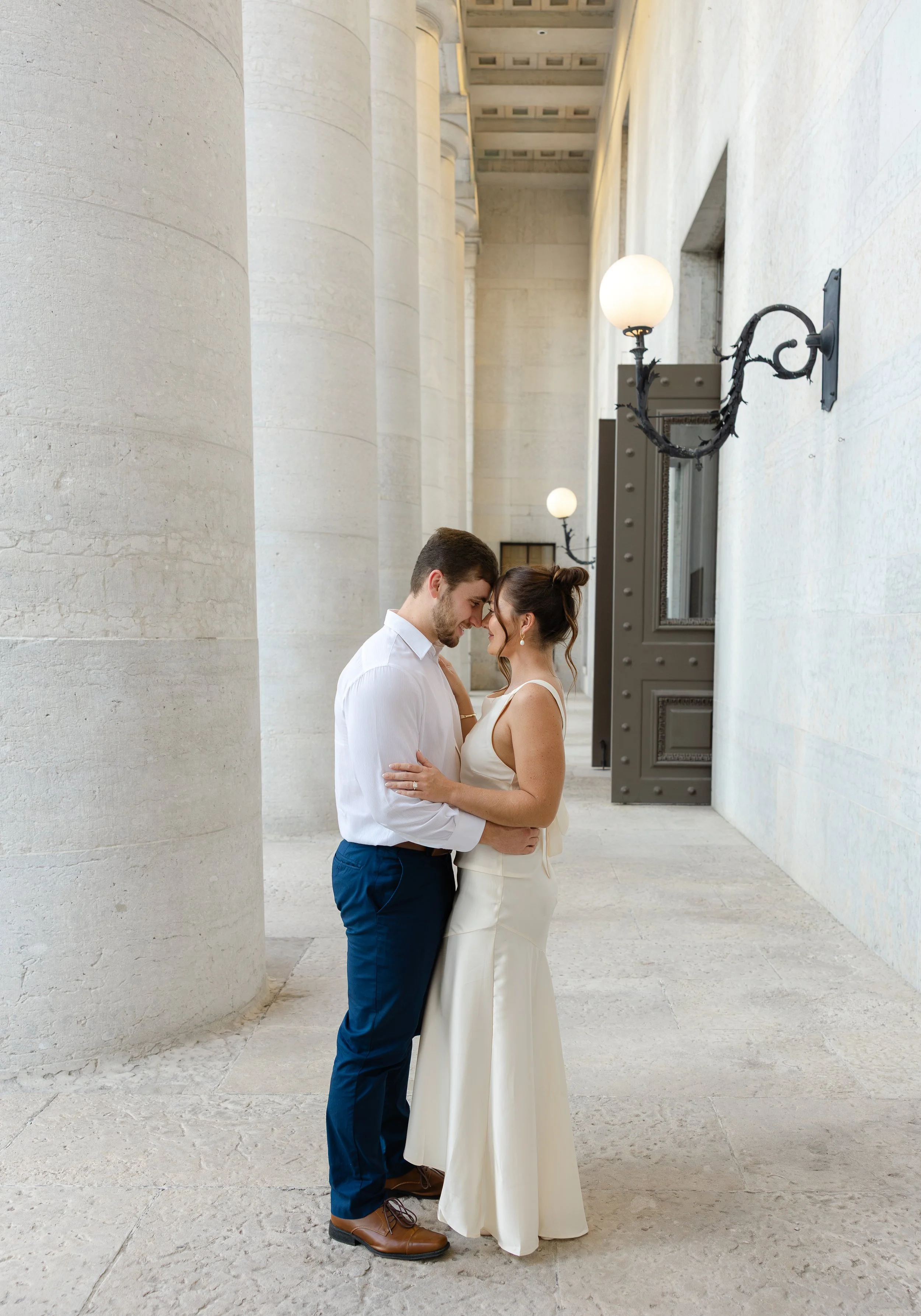 A couple in wedding attire standing close together, touching foreheads and smiling, in a grand, classical architectural setting with tall columns and wall-mounted lamps.