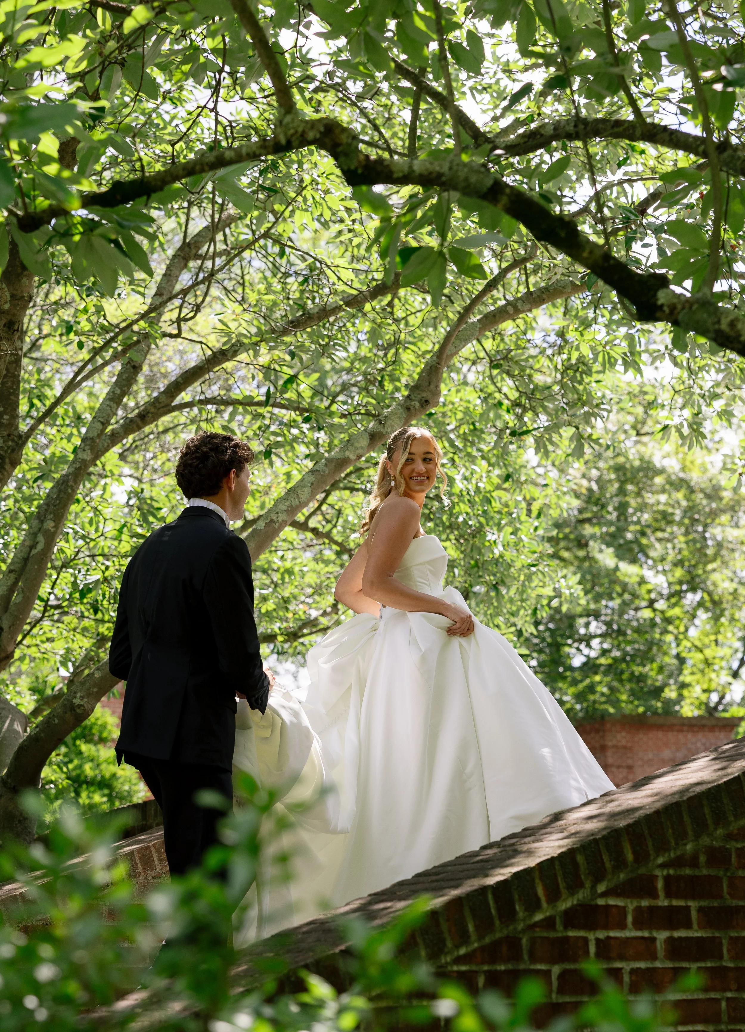 A bride in a white wedding gown standing on a brick wall under green trees, smiling, with a groom in a black suit holding her wedding dress.