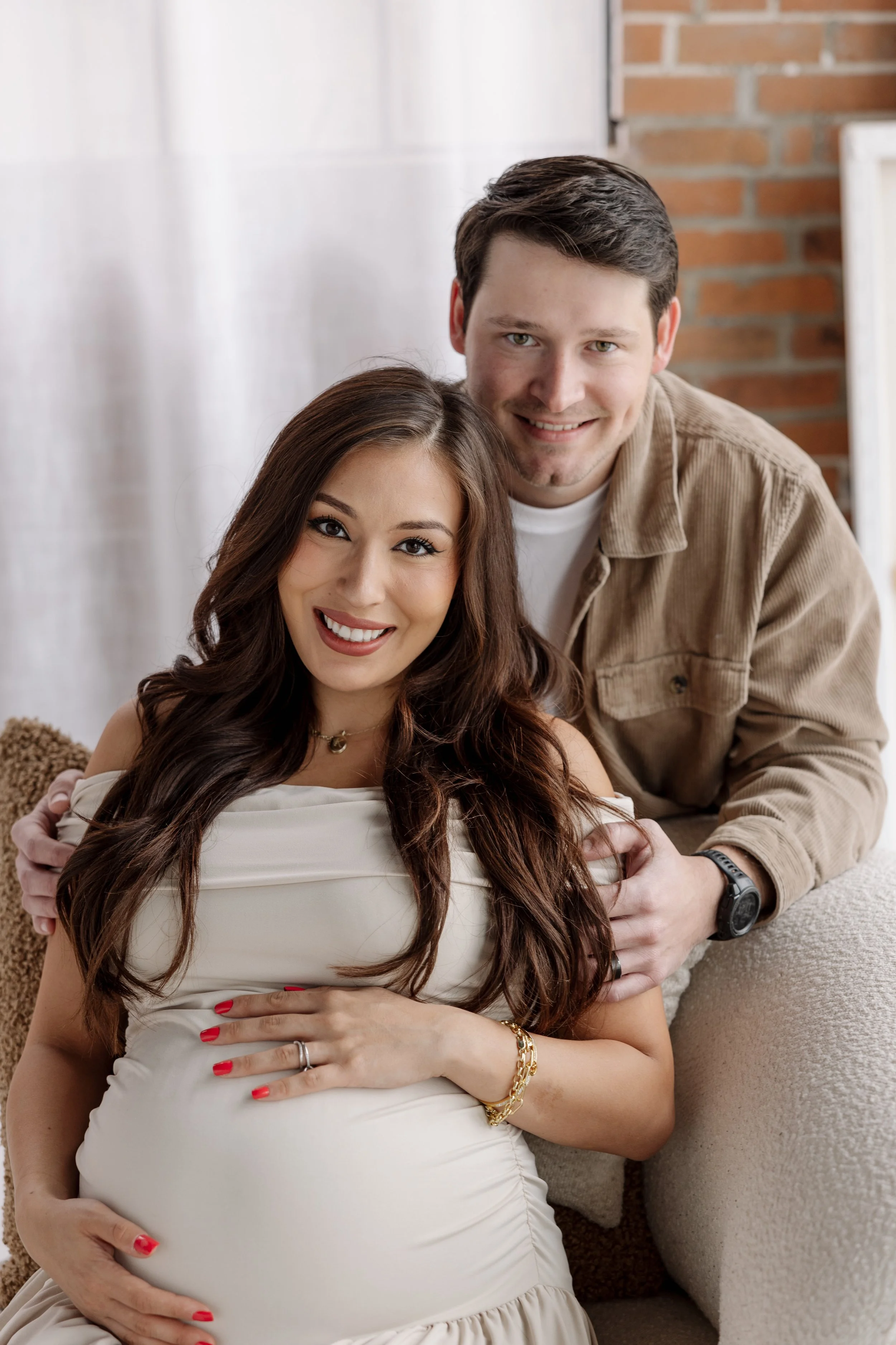 A pregnant woman with long brown hair and red nail polish smiling, with her partner behind her, in a cozy room with brick wall and white background.