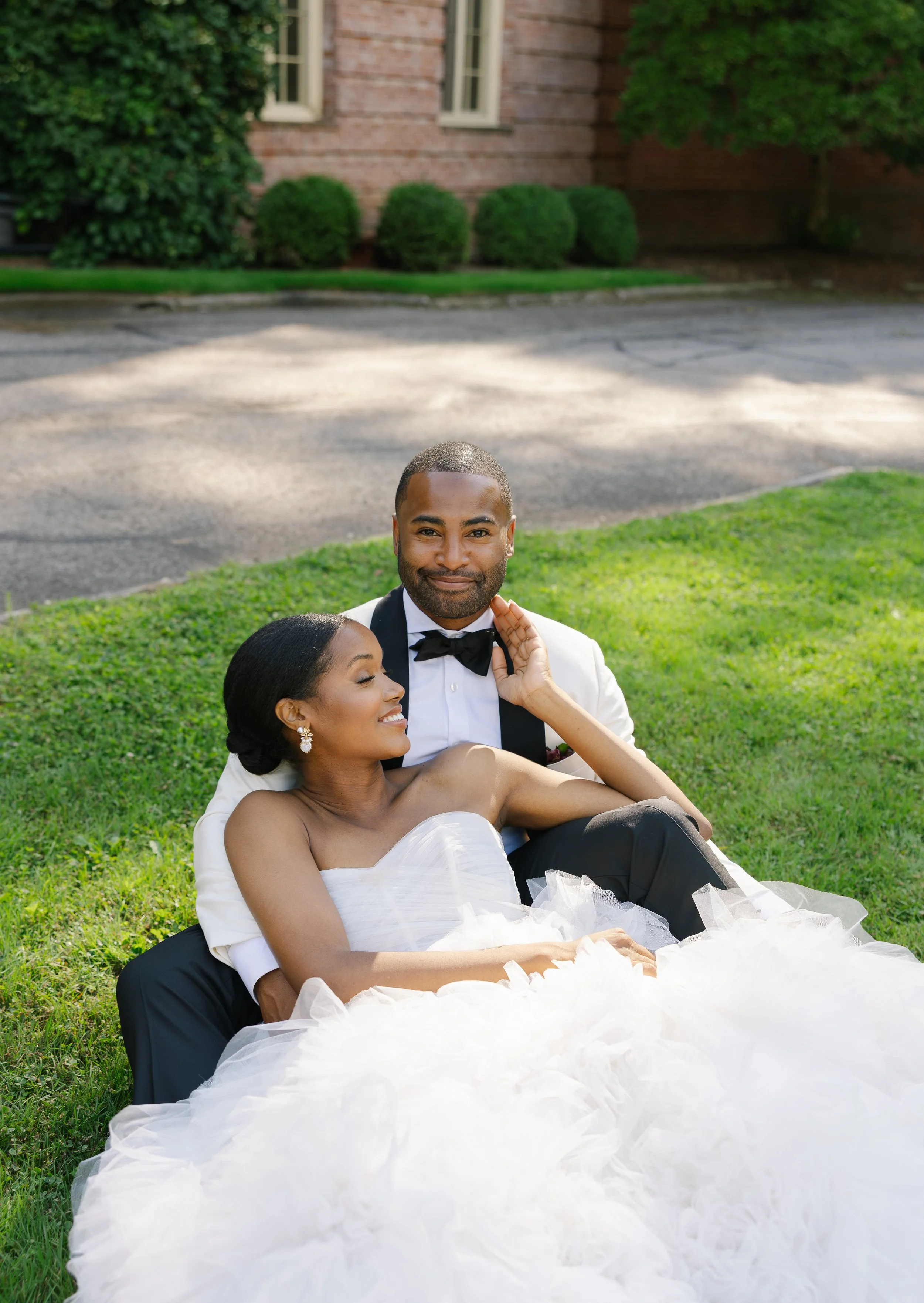 A newlywed couple sitting on the grass in front of a brick house, celebrating their wedding day. The bride is wearing a white wedding gown and the groom is in a white tuxedo with a black bow tie.