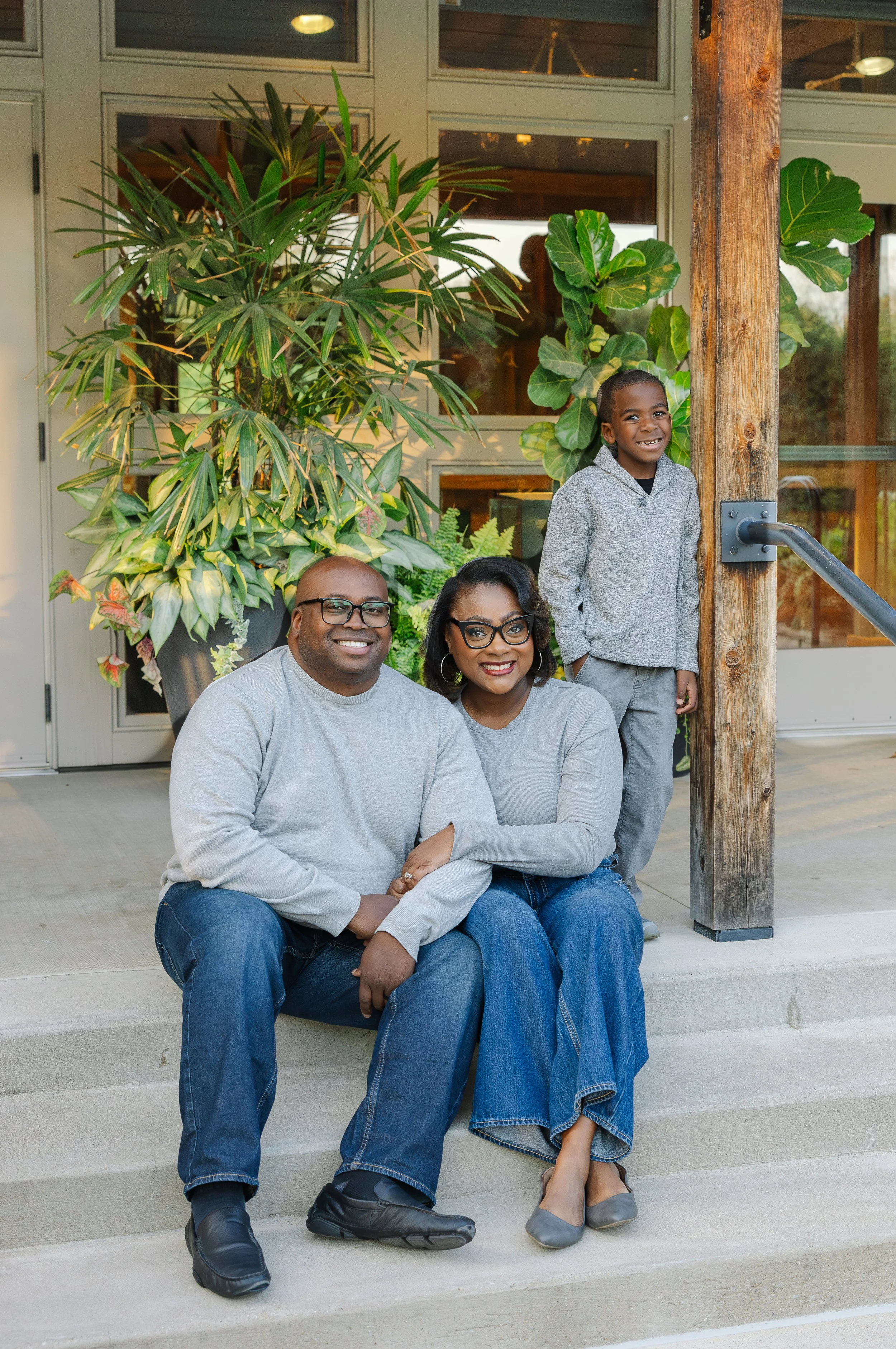 A happy family of three sitting on the steps outside a building with large potted plants in the background. The man and woman are seated closely, smiling at the camera, while the boy stands behind them, also smiling.
