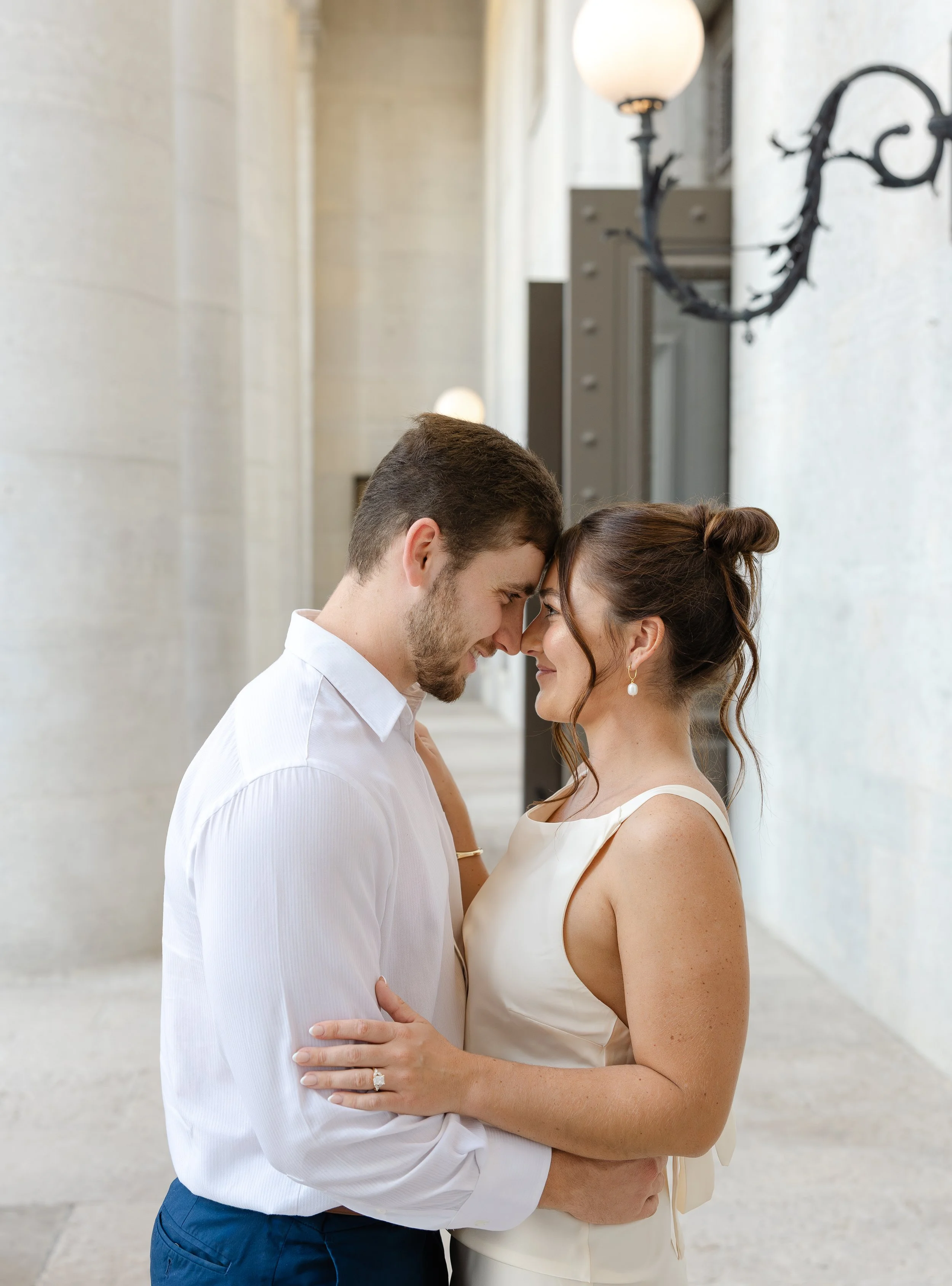 A couple sharing an intimate moment, faces touching, outdoors near a white wall with black decorative lighting fixtures.