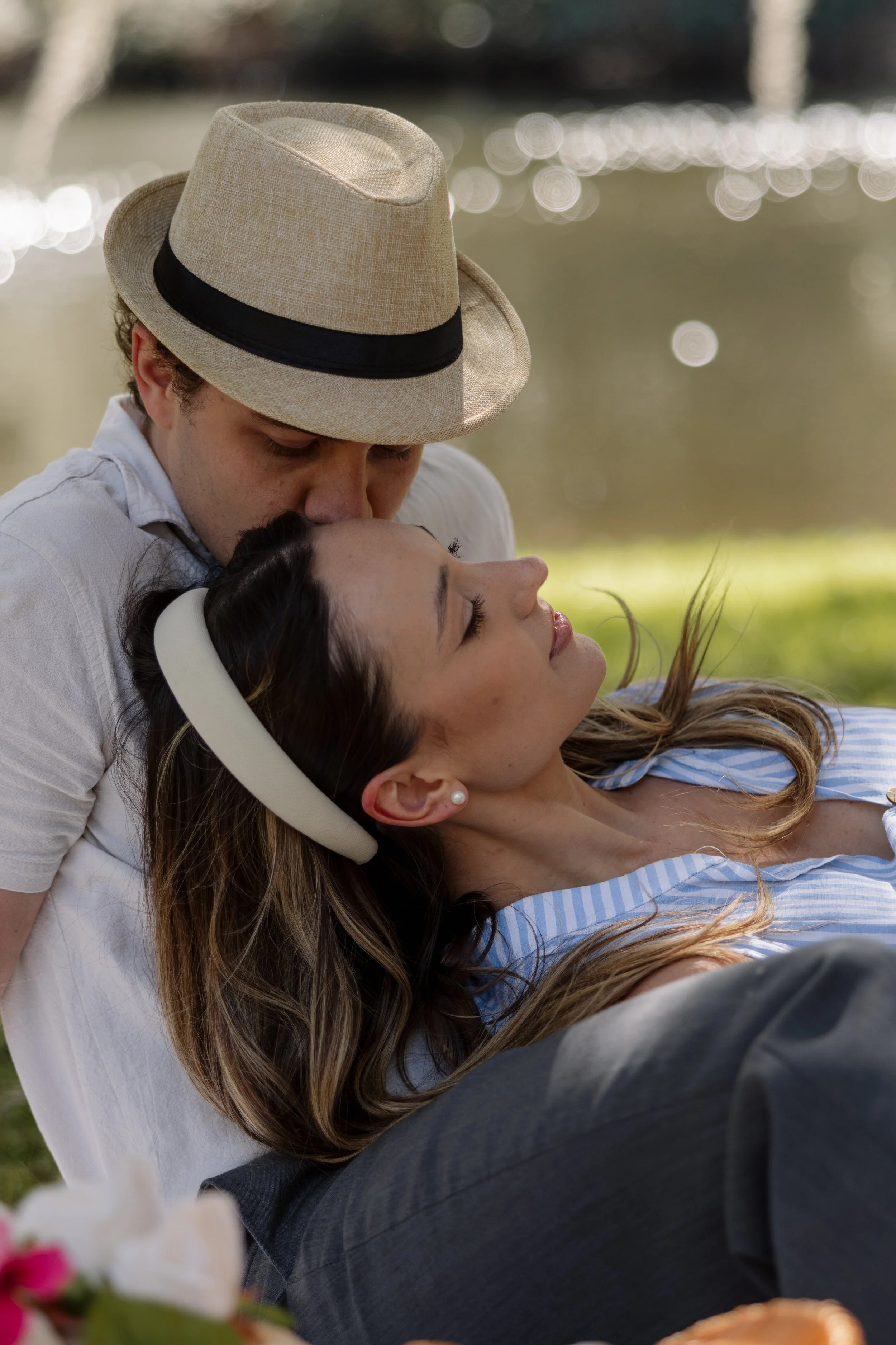 A man with a beige fedora hat kissing a woman with long brown hair and a white headband, lying outdoors on green grass near a body of water.