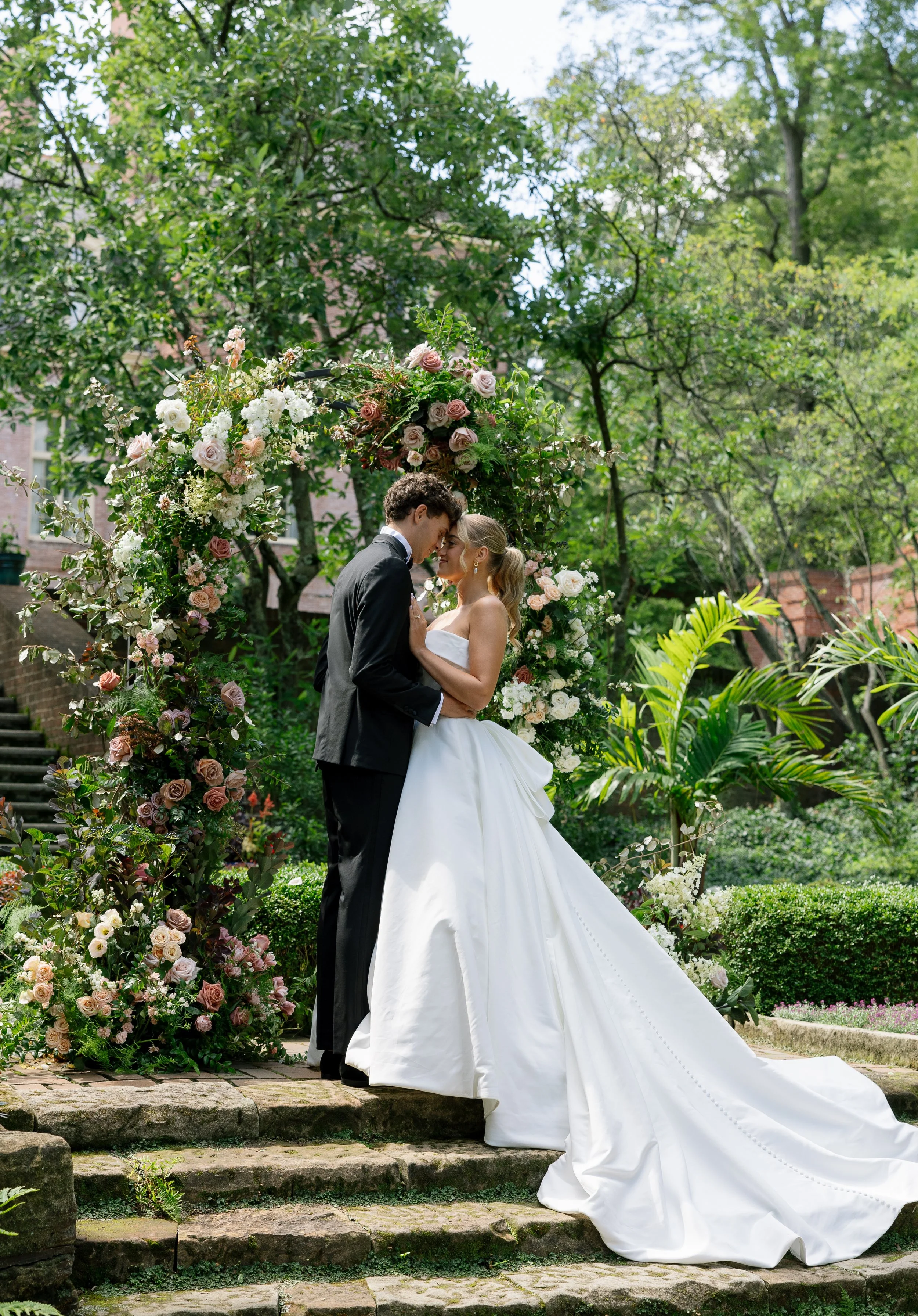 A bride and groom share a kiss beneath a floral arch in a lush garden, with green trees and plants in the background.