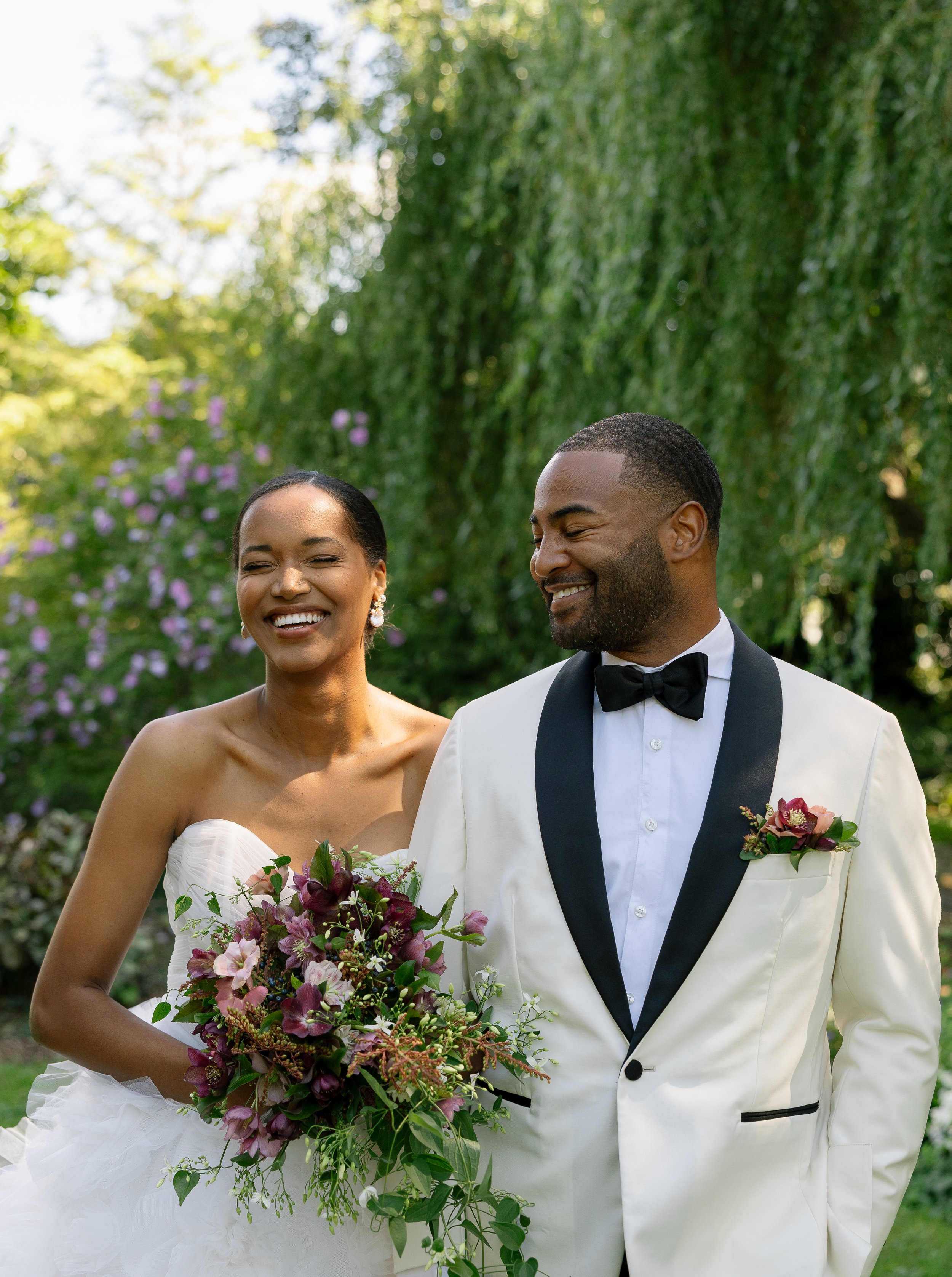 A happy bride and groom smile outdoors, dressed in wedding attire. The bride is holding a bouquet of purple and green flowers, and the groom wears a white tuxedo with black lapels and a black bow tie. There are green trees and purple flowers in the b